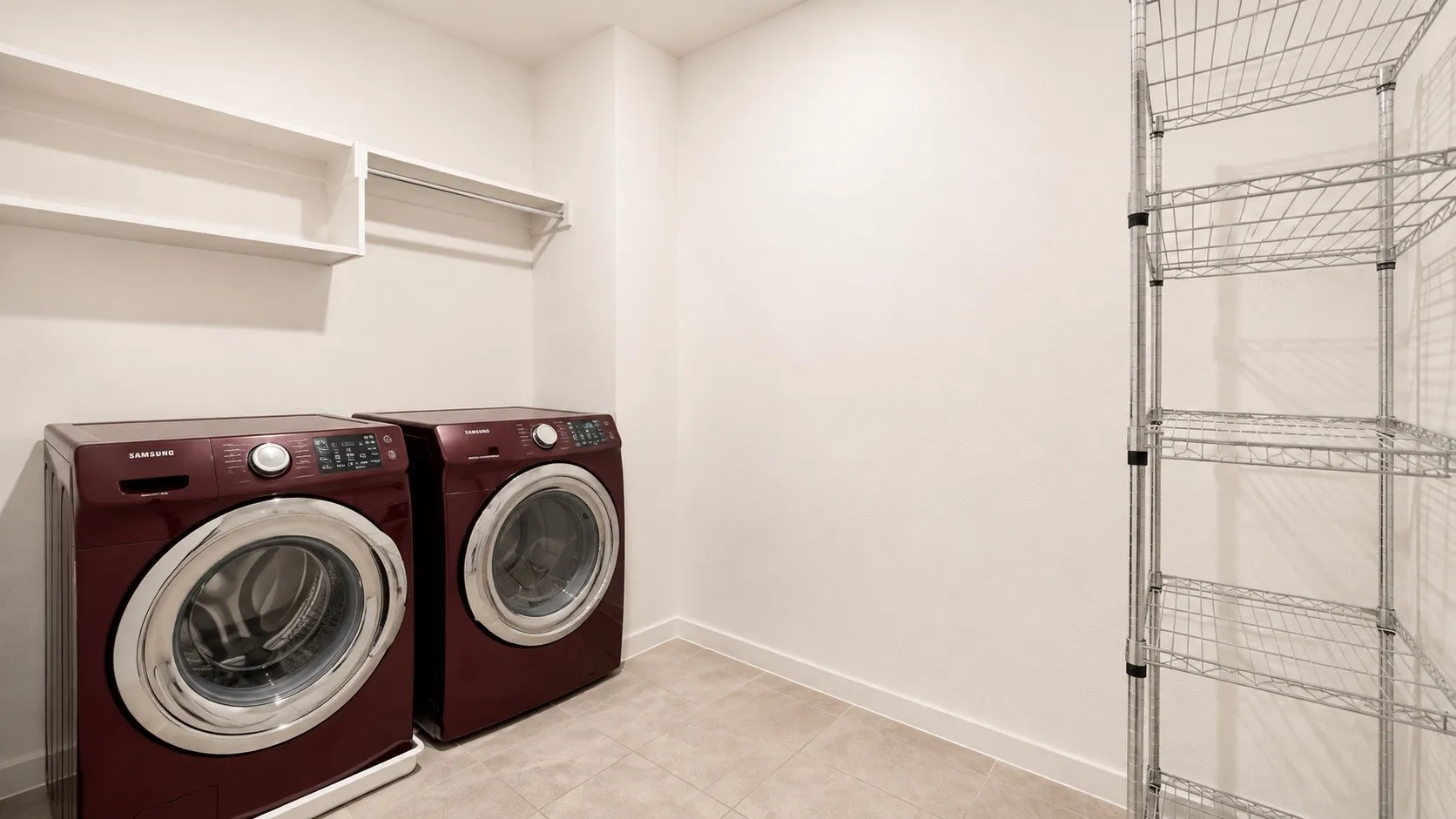 Dedicated laundry room featuring side-by-side front-load washers and dryers in burgundy finish, neutral tile flooring, white walls, floating shelving, and wire storage racks for organized utility storage