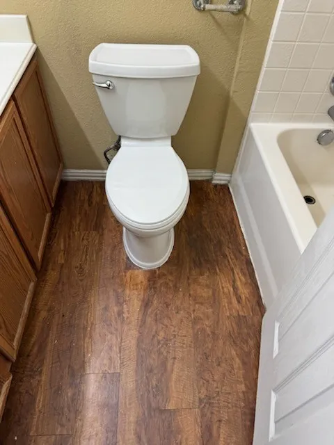 Wood-finish flooring with a white toilet, adjacent to a white bathtub with tiled surround