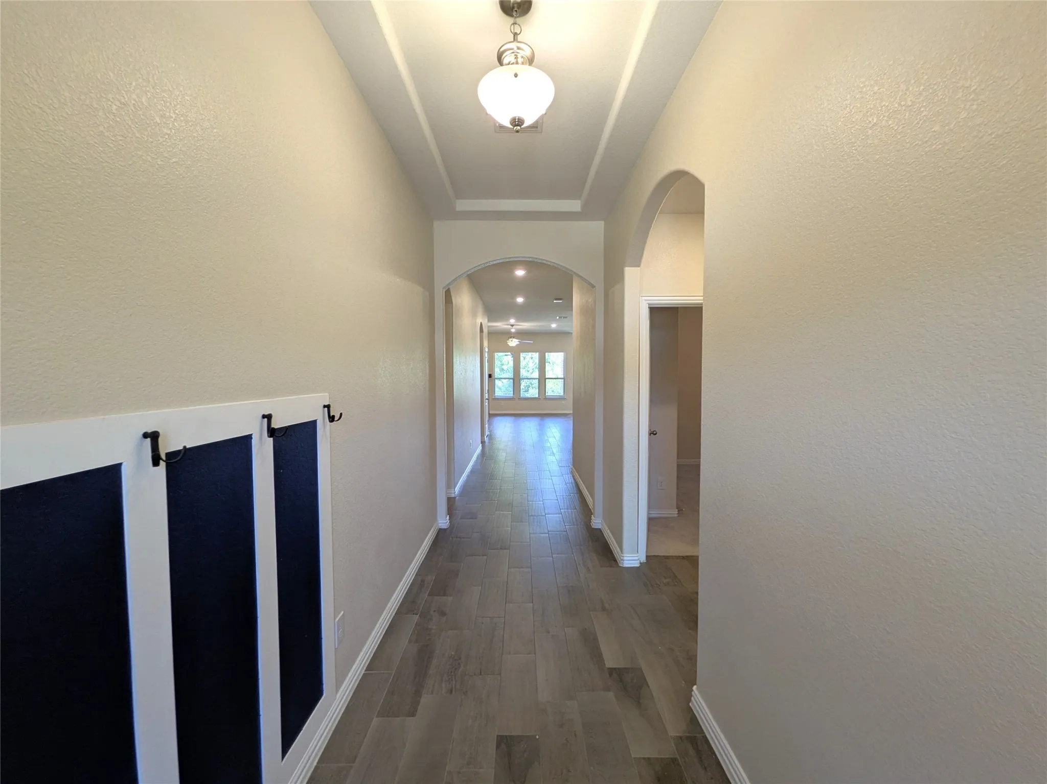 Elegant hallway featuring crisp white walls, coffered ceiling detail with pendant lighting, wide-plank wood-finish flooring in a warm gray tone, and arched architectural elements