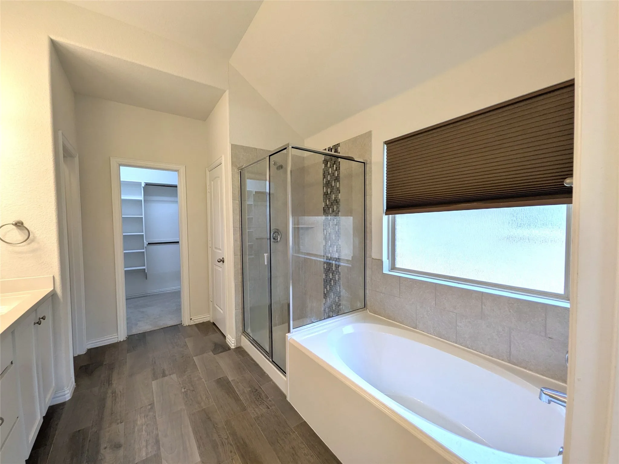 Spa-inspired master bath featuring a soaking tub with tile surround, a frameless glass-enclosed shower with decorative tile accent wall, dual vanity with white cabinetry, wood-finish flooring, a walk-in closet, and a large window with motorized cellular shade for natural light and privacy