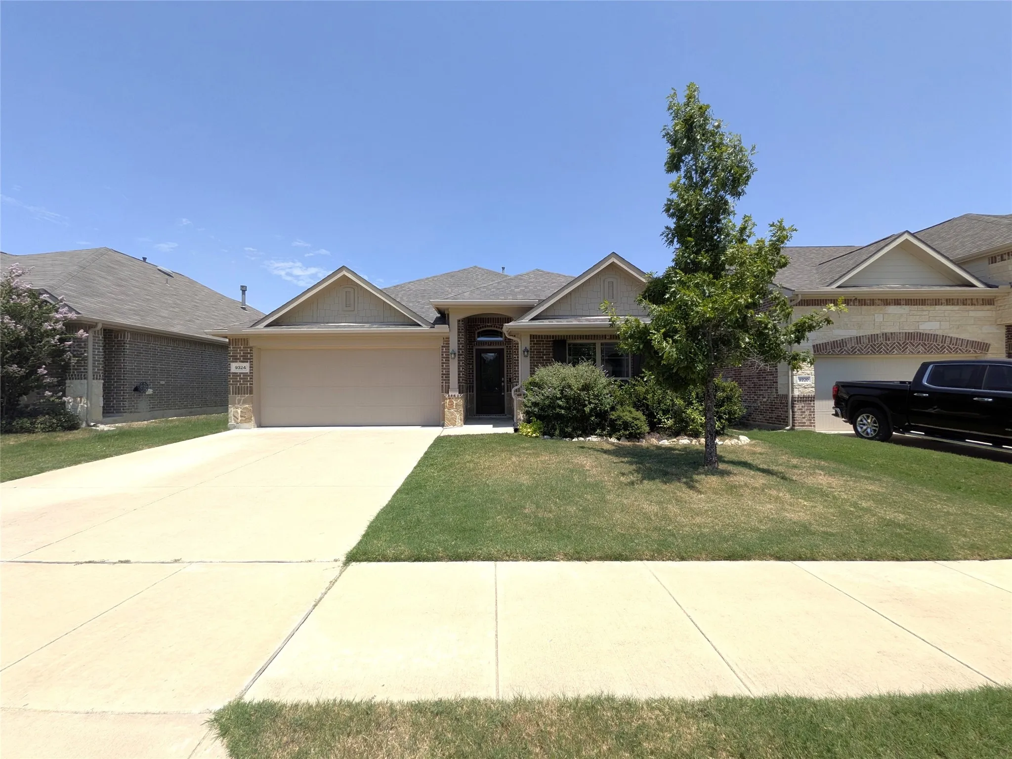 Welcoming single-story home featuring a spacious driveway, neutral exterior with brick and stone accents, and mature landscaping including established trees