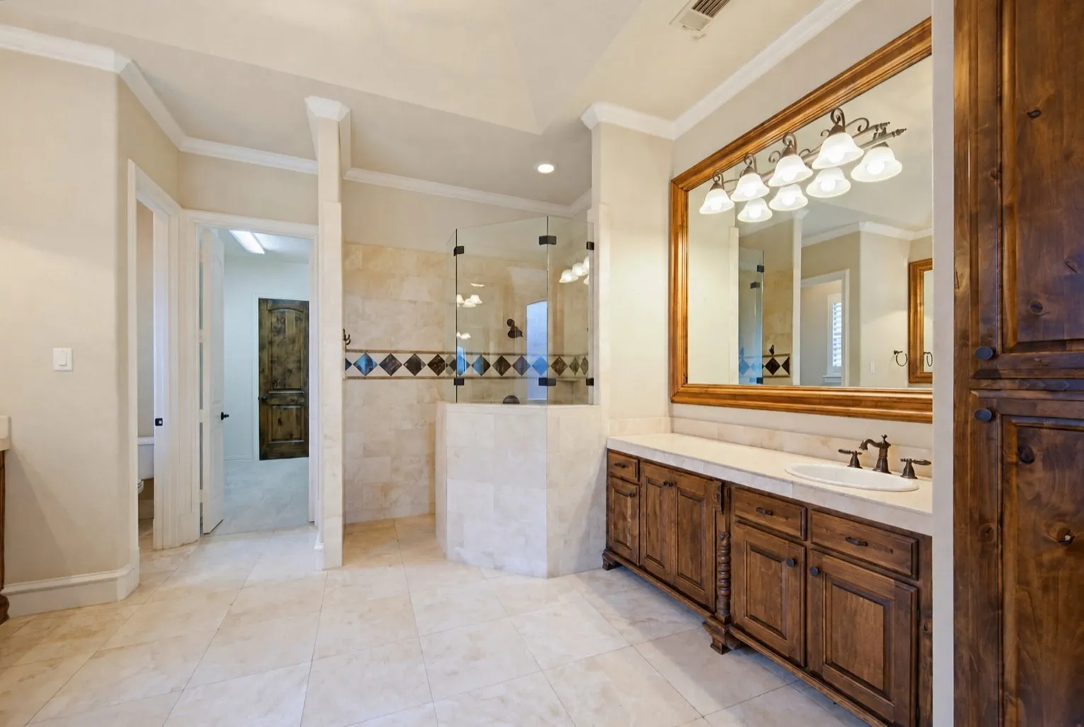 Spa-inspired primary bathroom featuring a spacious walk-in shower with decorative tile accents, dual vanity with wood cabinetry and framed mirror, recessed lighting, and travertine-finish flooring throughout