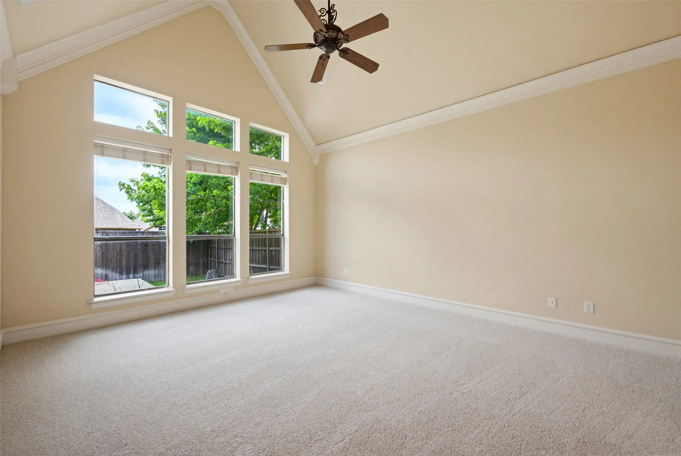 Bright and spacious primary bedroom showcasing soaring vaulted ceilings with crown molding, large multi-pane windows flooding the space with natural light, neutral carpet flooring, ceiling fan with dark finish, and white trim throughout