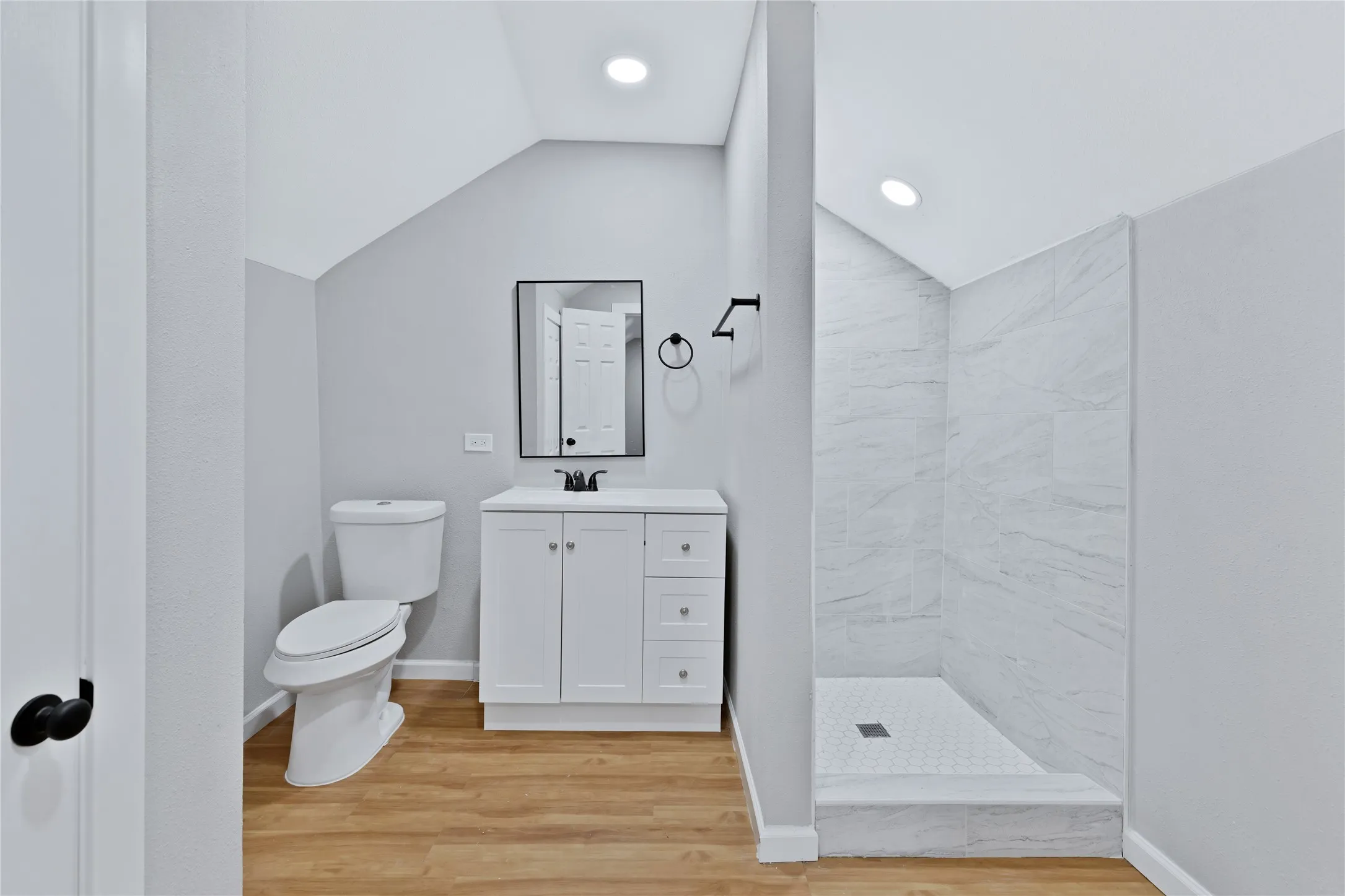 Modern bathroom with wood-finish flooring, white vanity, and a walk-in shower featuring light-toned tile walls and a white hex-tile floor
