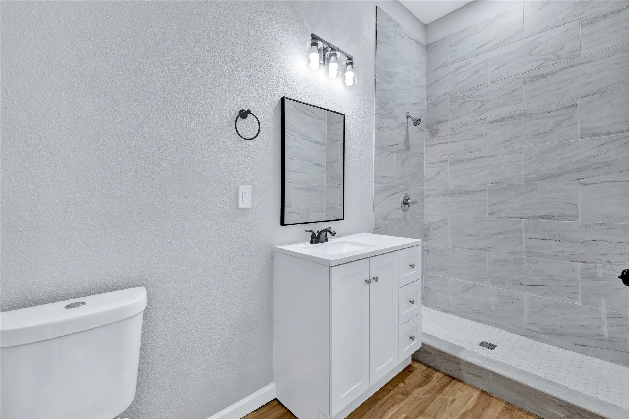 White vanity with integrated sink, black framed mirror, three-light vanity fixture, and a tiled walk-in shower with a mosaic floor