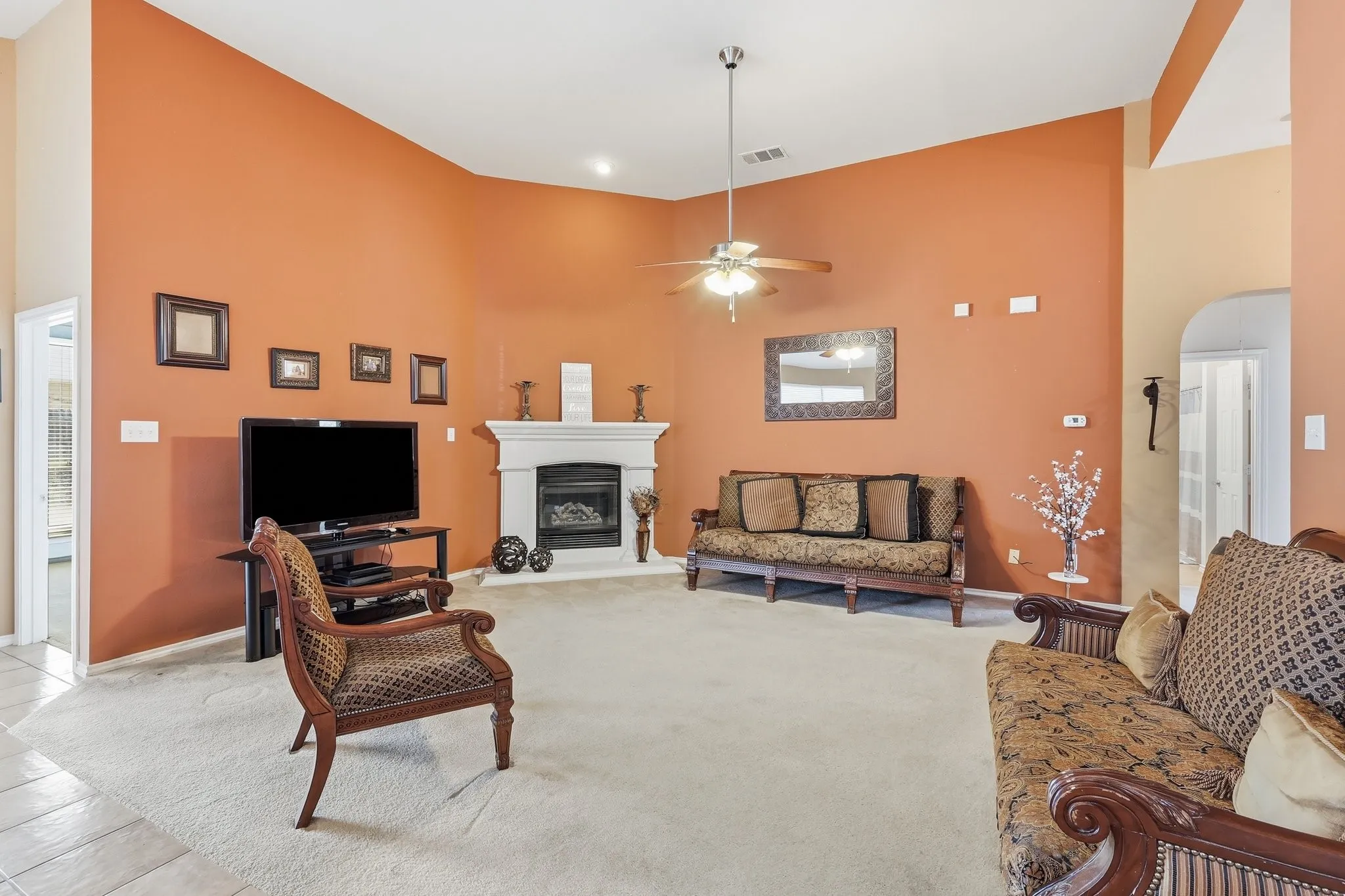 Spacious living area featuring a fireplace with a white mantel, ceiling fan, carpet flooring, and a tiled entryway