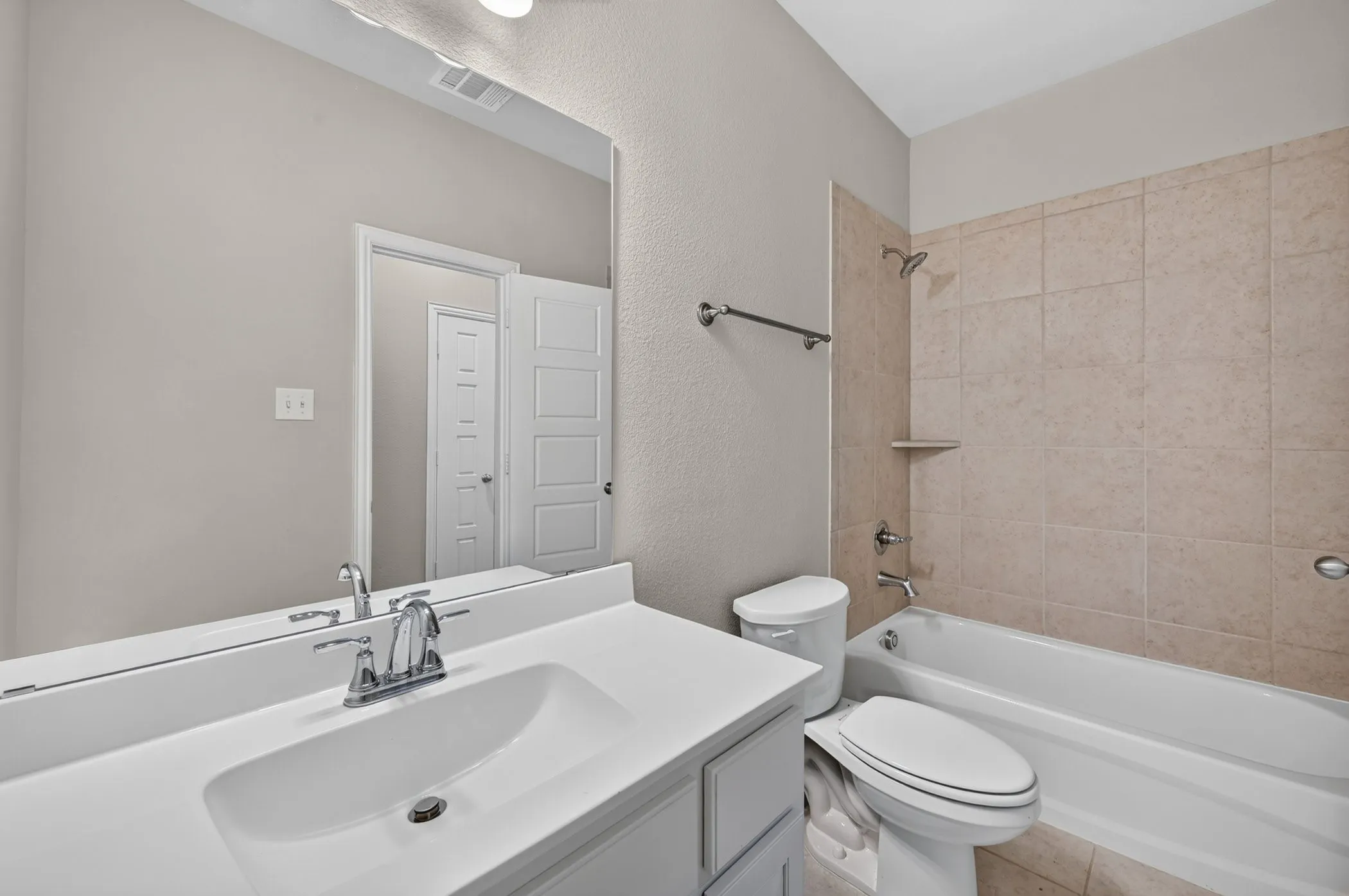 Bathroom featuring a white vanity with integrated sink, chrome faucet, large frameless mirror, and a combination tub/shower with tan tiled surround