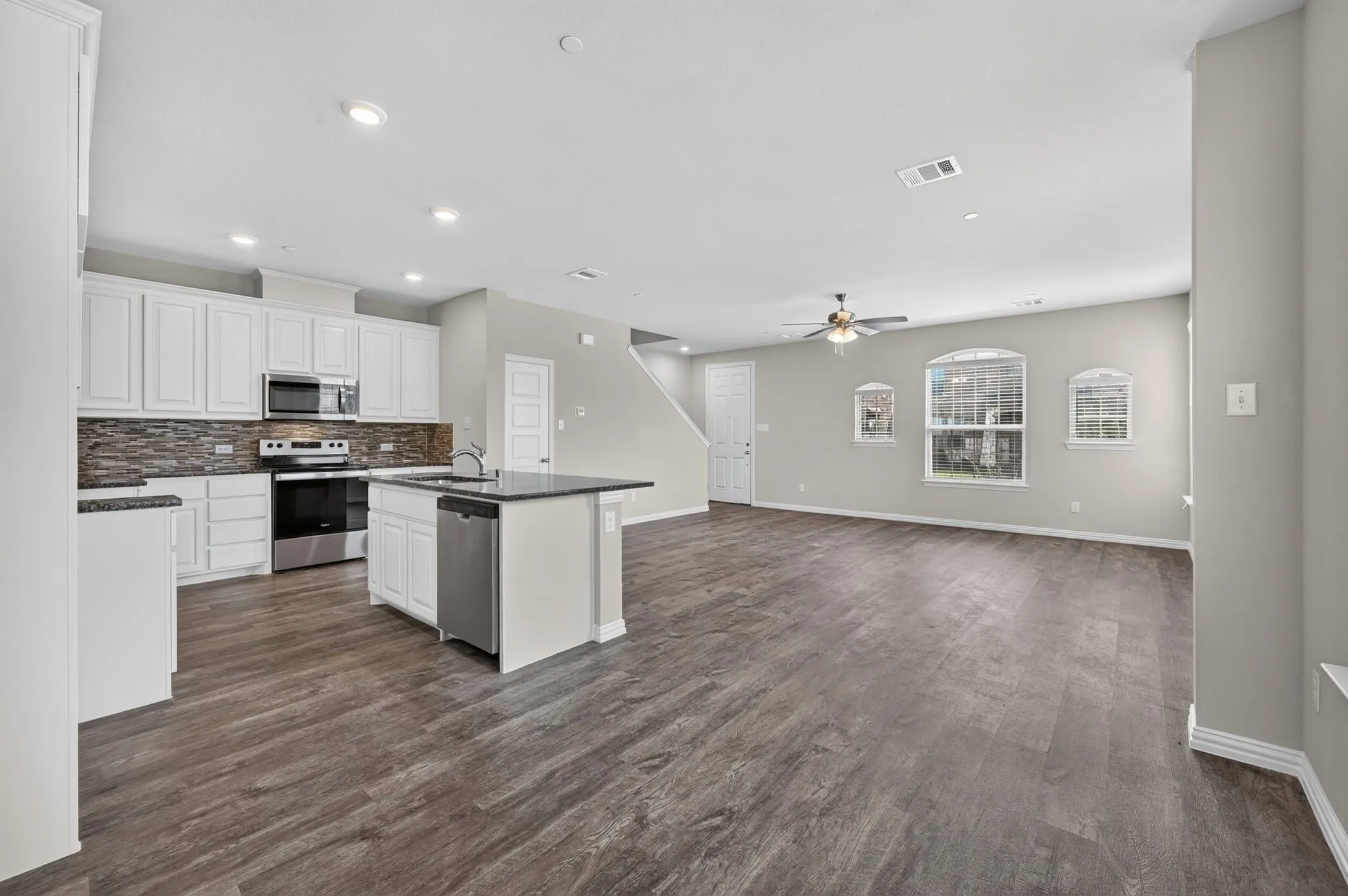 Open-concept living space featuring wood-finish flooring, a kitchen island with sink, stainless steel appliances, white cabinetry, and a stacked stone backsplash
