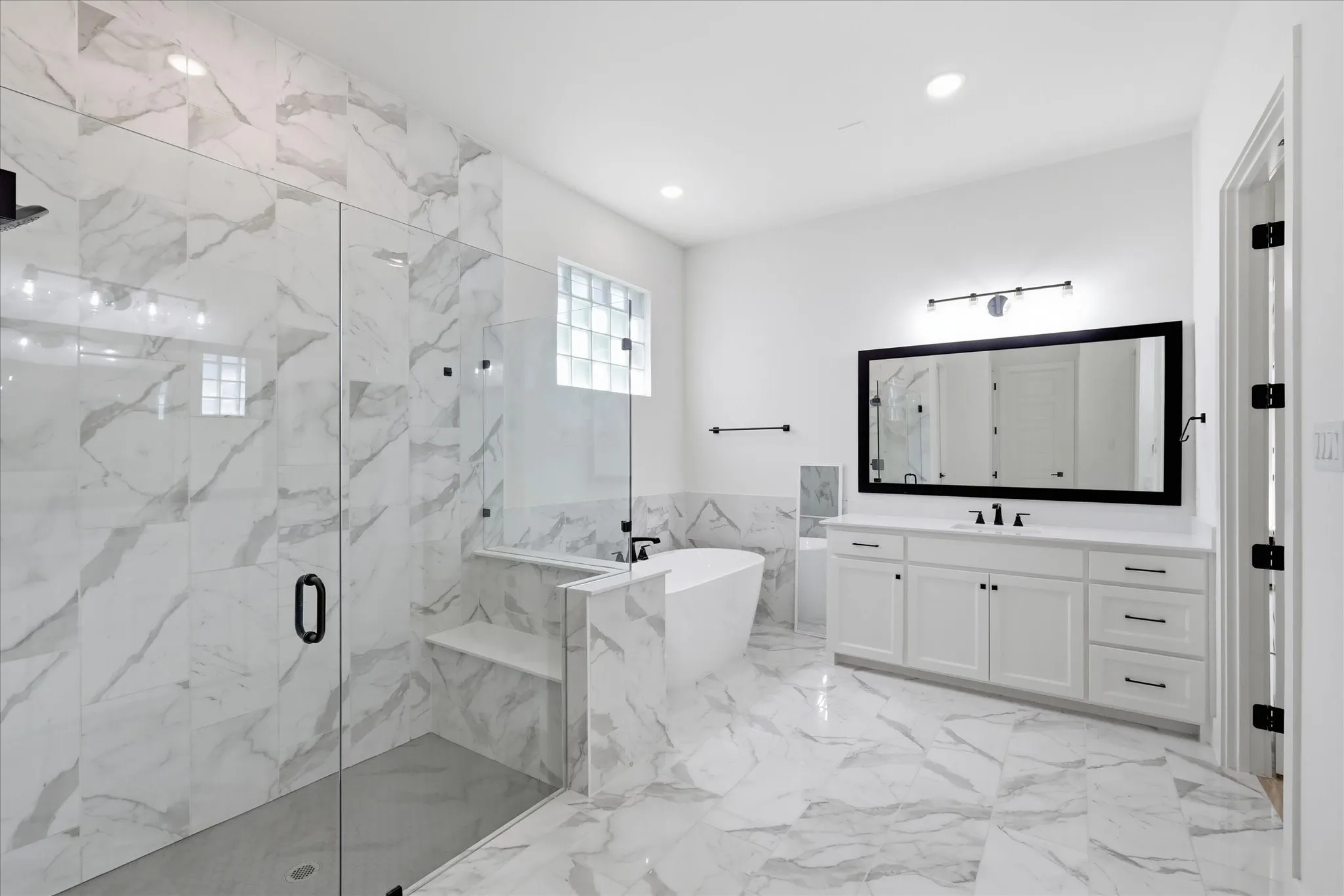 Spacious bathroom featuring large format marble-look tile flooring, a glass-enclosed shower with a built-in bench, a freestanding soaking tub, and a dual vanity with a large framed mirror