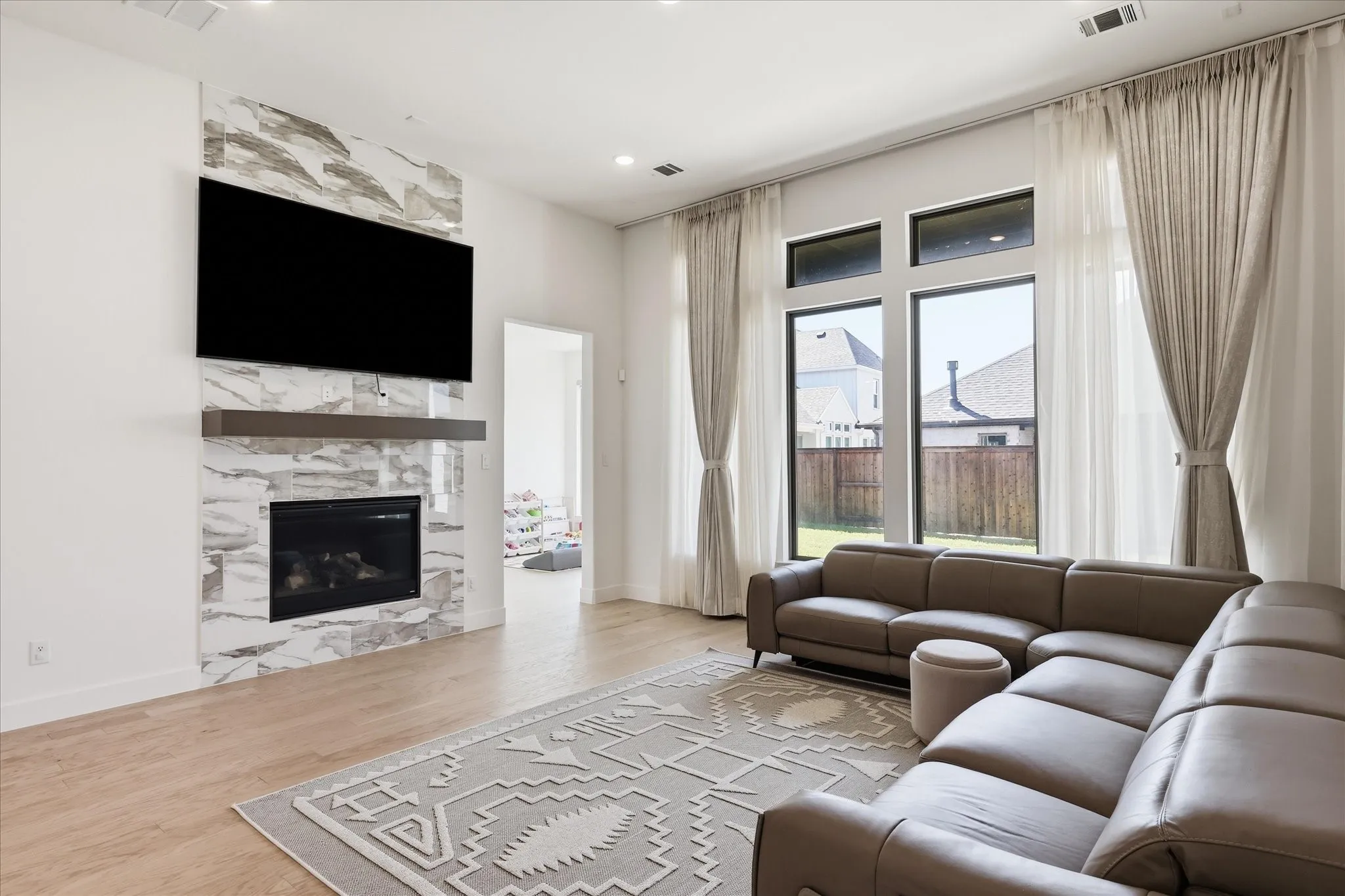 Sophisticated living room showcasing a linear gas fireplace with marble surround and floating television mount, wide-plank wood-finish flooring, floor-to-ceiling windows with operable glass doors, and custom drapery hardware with neutral fabric panels