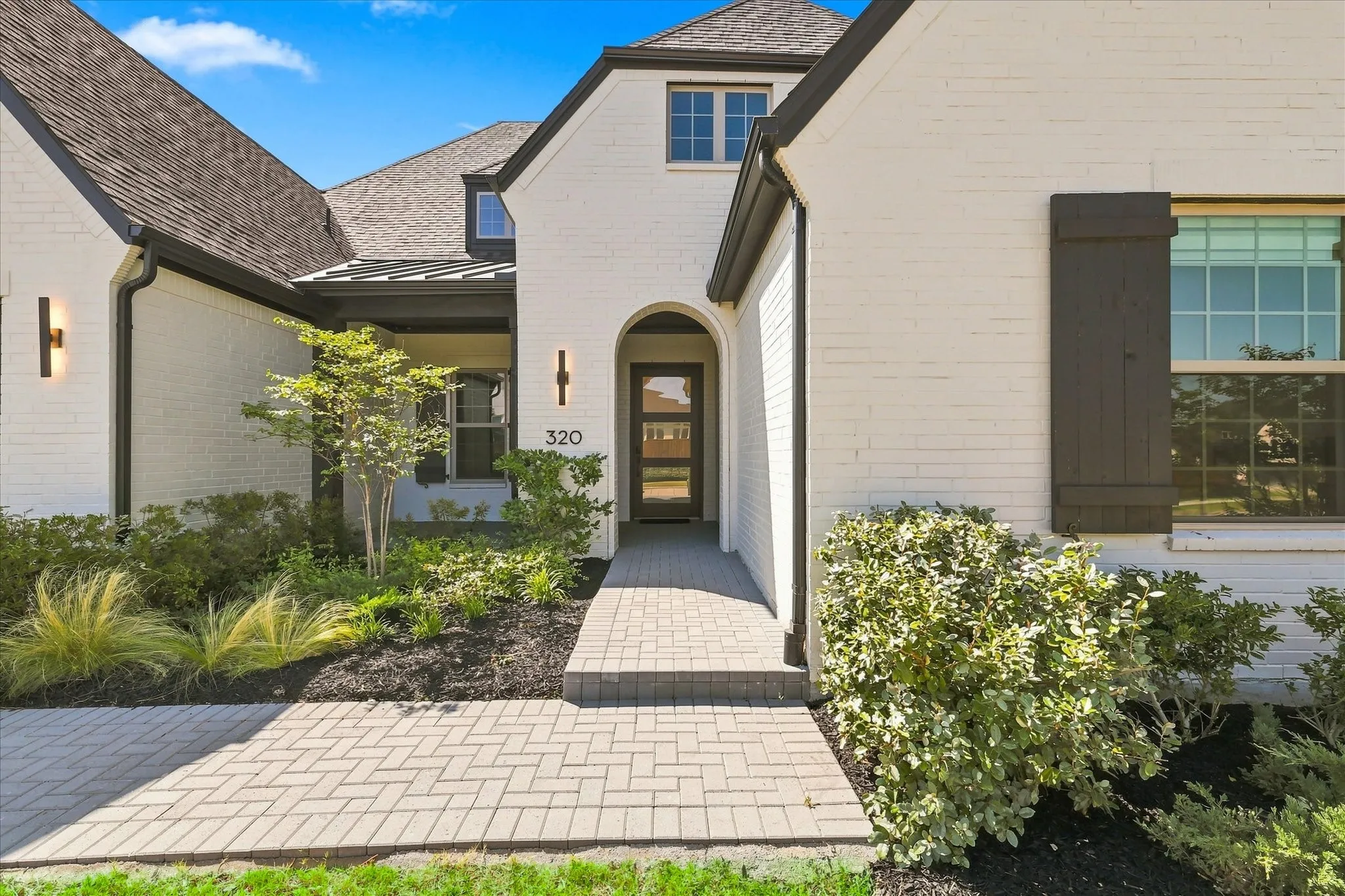 Welcoming entry courtyard featuring permeable paver hardscaping, mature landscaping with ornamental grasses and shrubs, and a charming arched wooden entry door