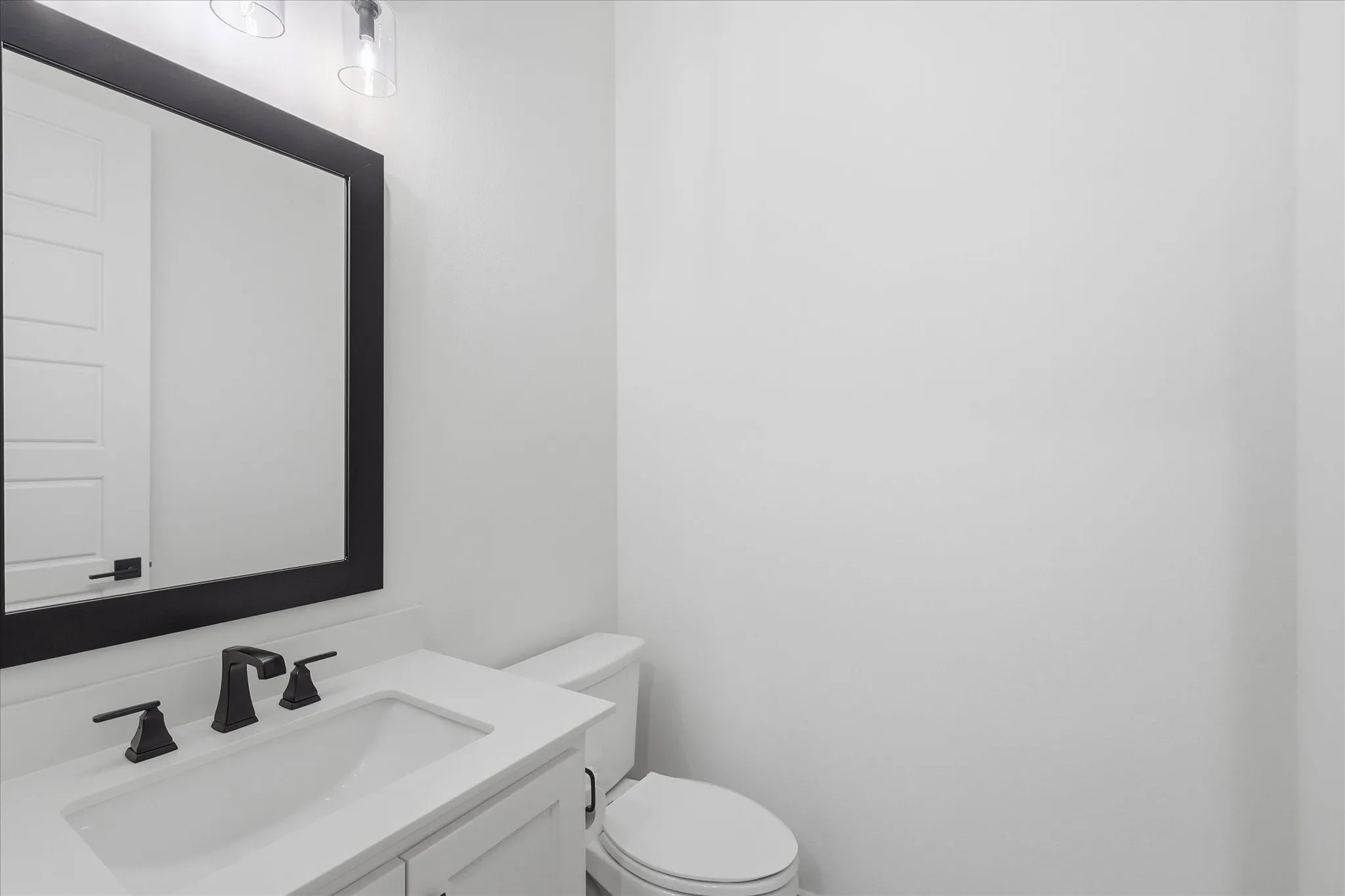 Clean and contemporary bathroom featuring a white porcelain sink with chrome fixtures, large framed mirror with black trim, white ceramic toilet, and bright white wall surfaces throughout
