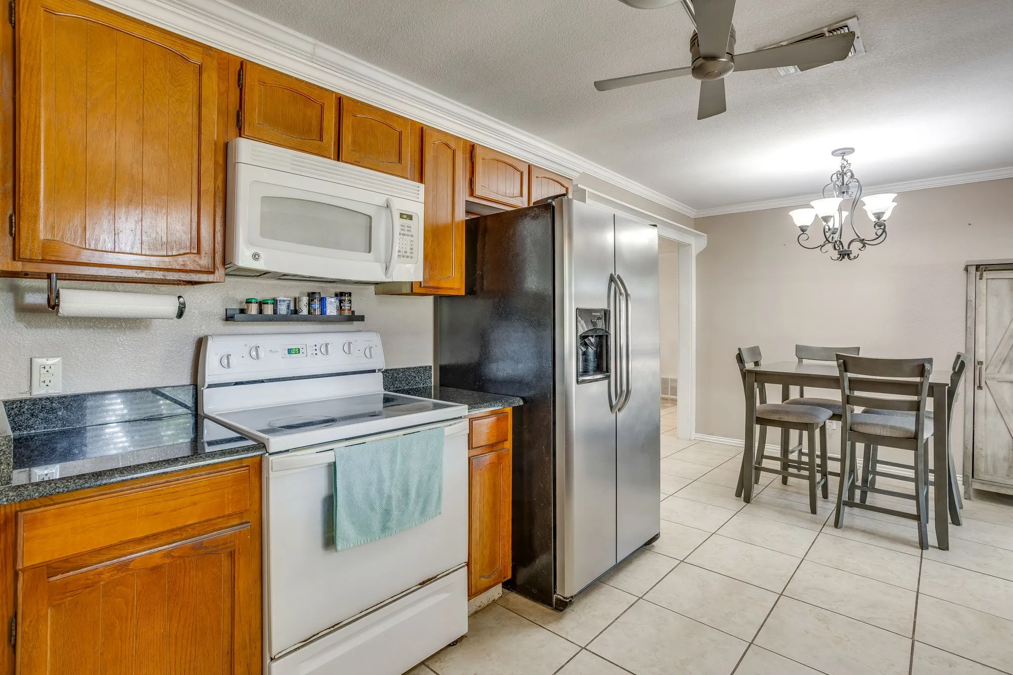 Spacious kitchen featuring warm wood cabinetry, stainless steel appliances including side-by-side refrigerator, electric range, and microwave, dark countertops, and ceramic tile flooring