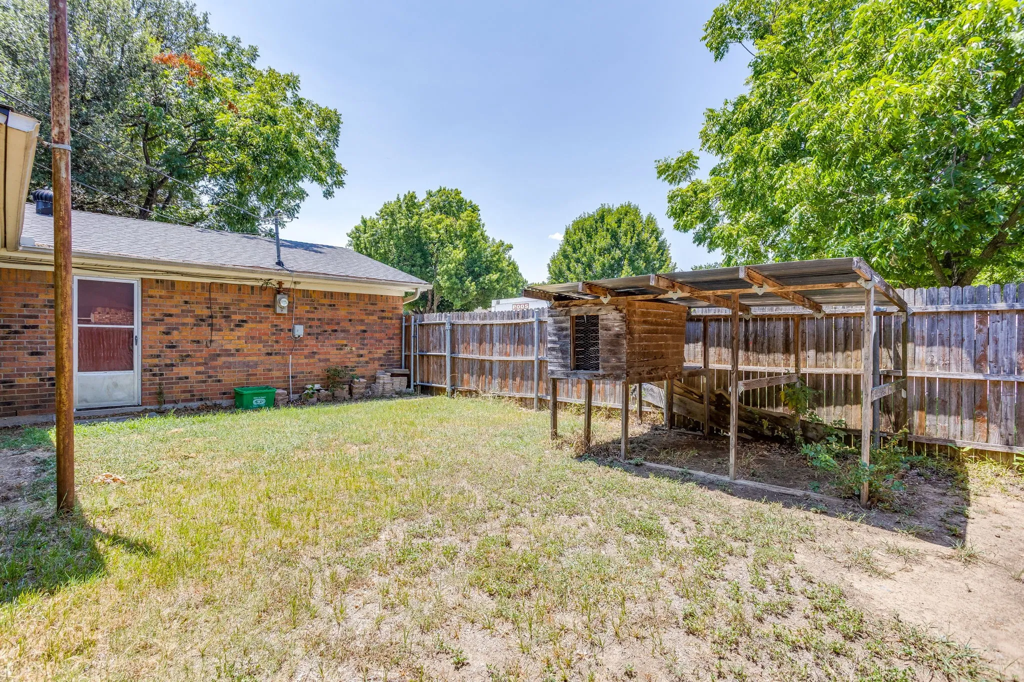 Expansive backyard featuring a partial brick exterior, a wood privacy fence, and mature trees providing natural shade