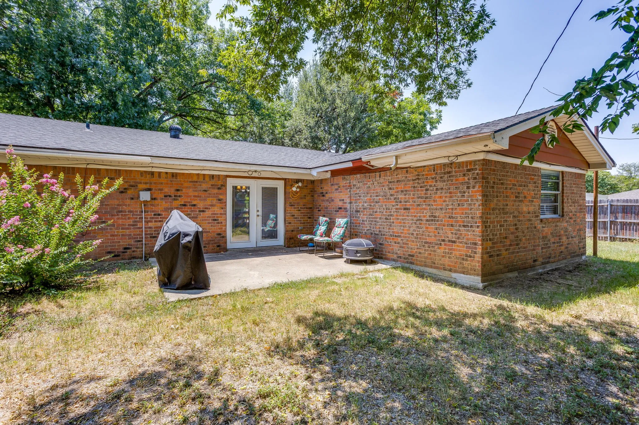 Brick ranch home featuring a covered patio with French doors, mature landscaping with established trees, and a spacious yard with natural lawn