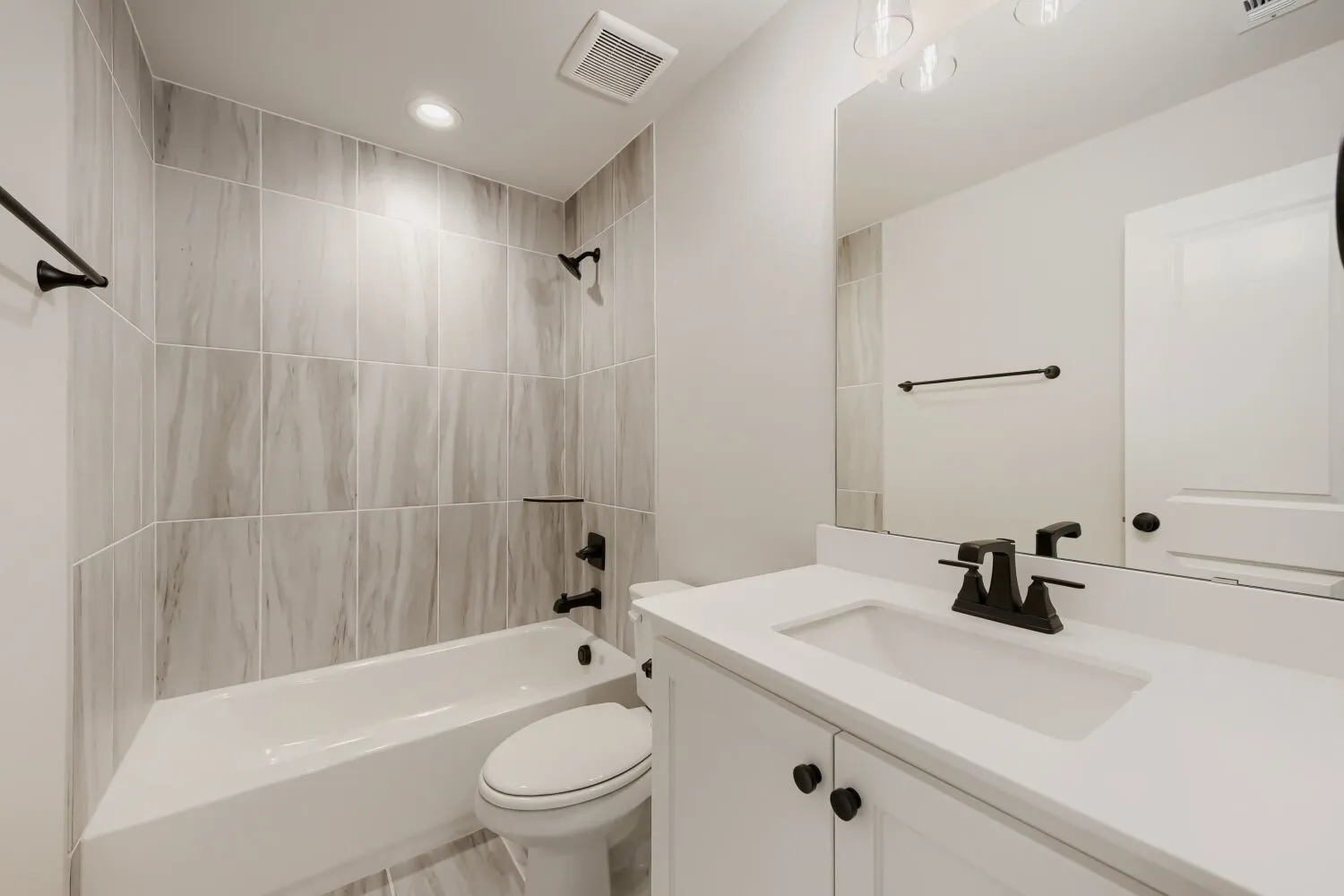 Bathtub with light-toned tiled surround, white vanity with integrated sink, white countertop, and dark-toned fixtures