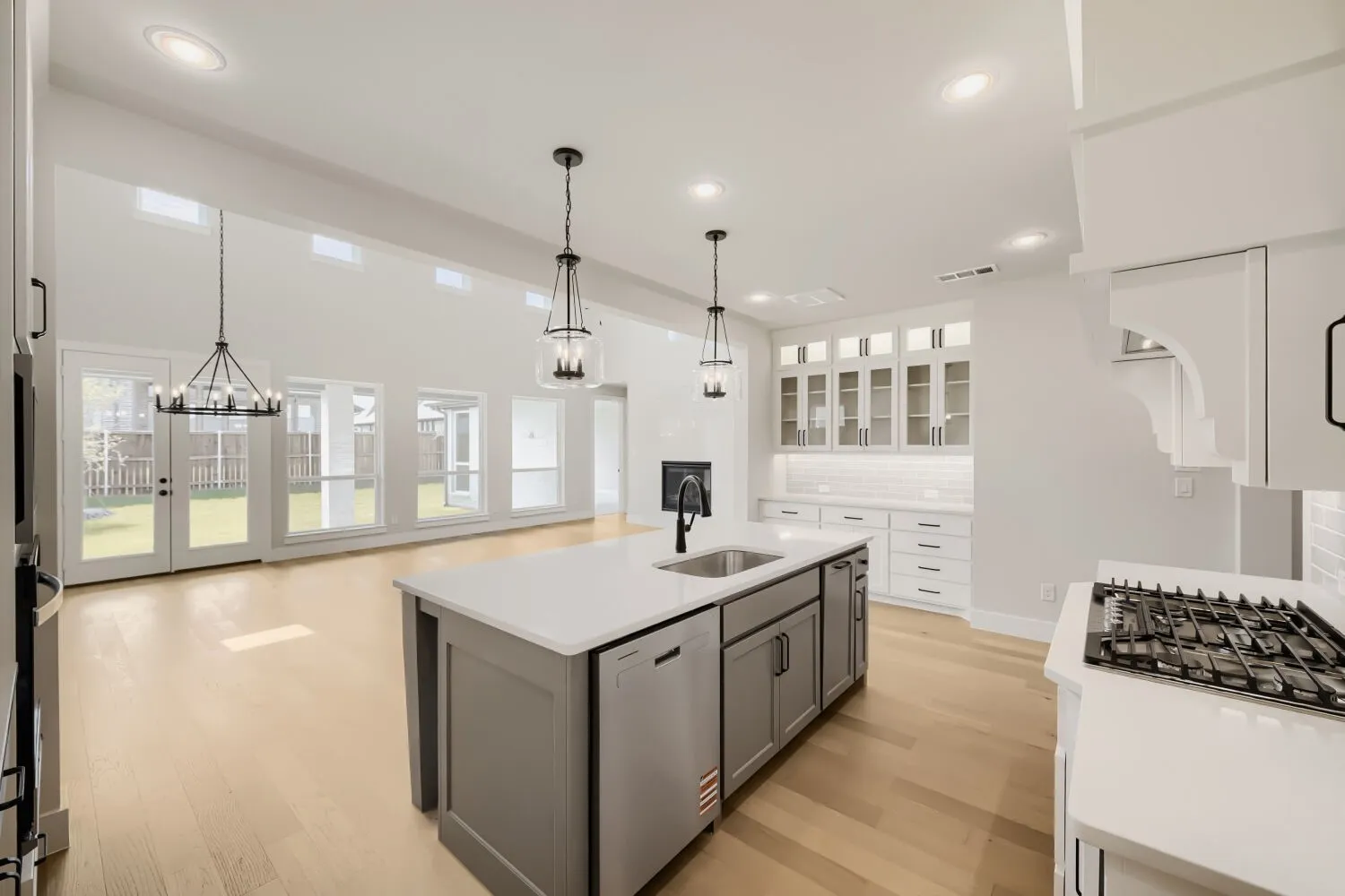 Open-concept kitchen and living area featuring a large center island with integrated sink, white subway tile backsplash, light wood-finish flooring, and a bank of glass-paneled cabinetry