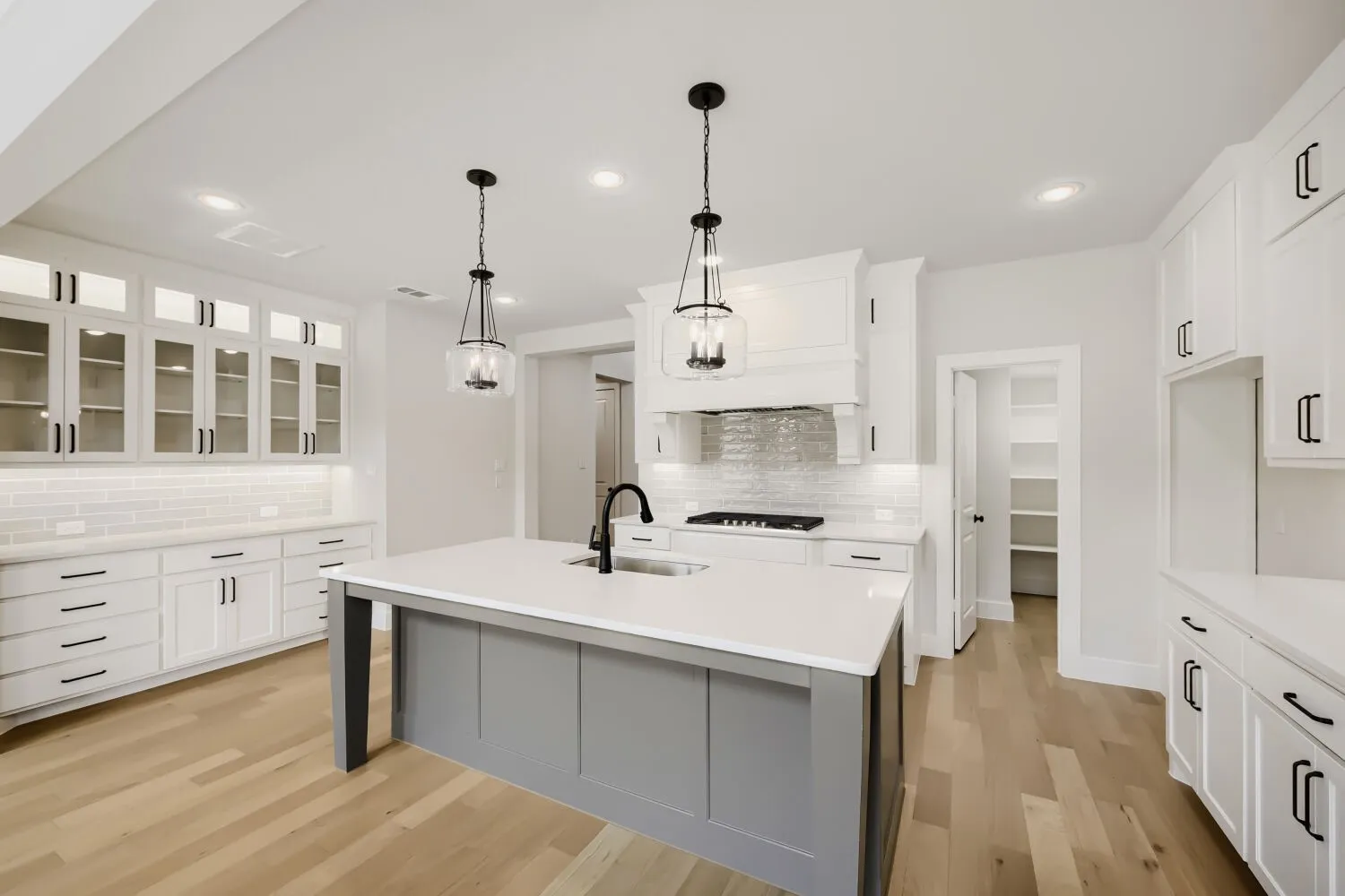 Kitchen featuring a central island with a sink, white cabinetry with dark hardware, wood-finish flooring, recessed lighting, and a pantry with built-in shelving