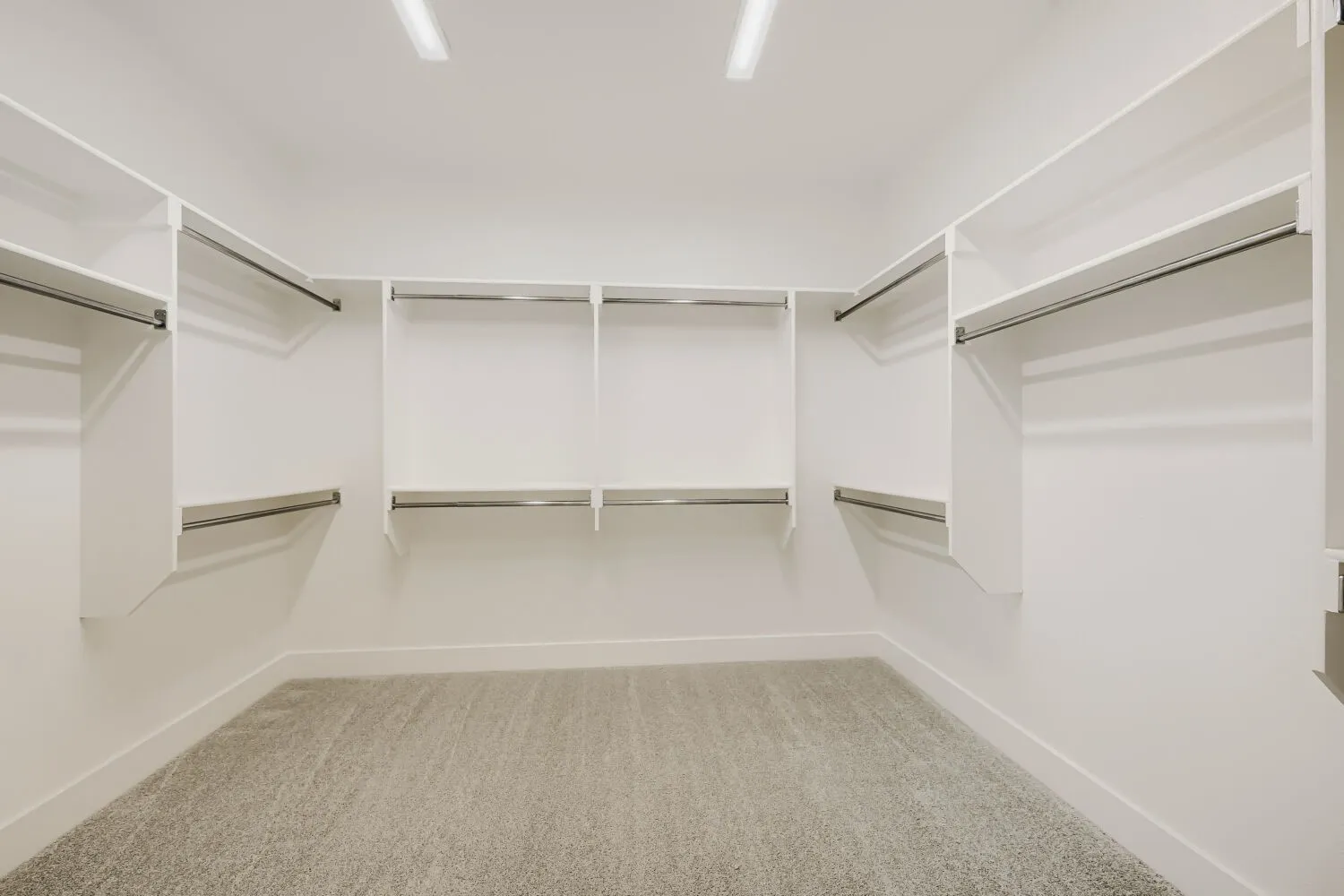 Recessed overhead lighting illuminating built-in white shelving, chrome hanging rods, and light gray carpeting