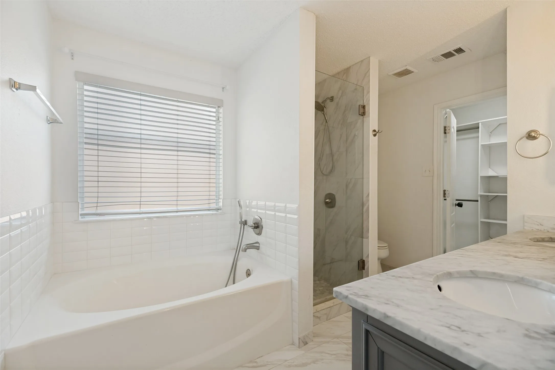 Soaking tub with white subway tile surround, glass-enclosed shower with tile walls, single vanity with integrated sink and stone countertop, and built-in closet shelving