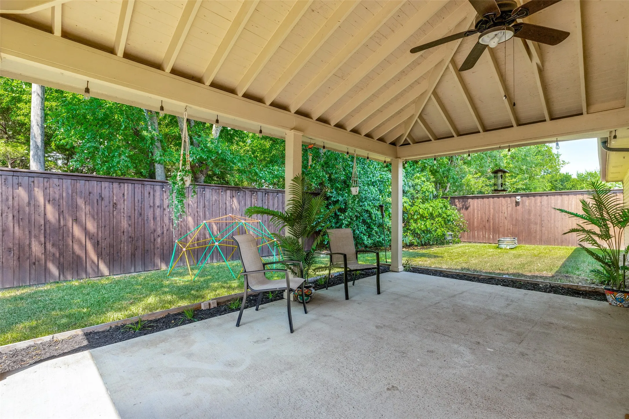 Covered patio featuring a ceiling fan, string lighting, and direct access to a fenced yard with mature trees