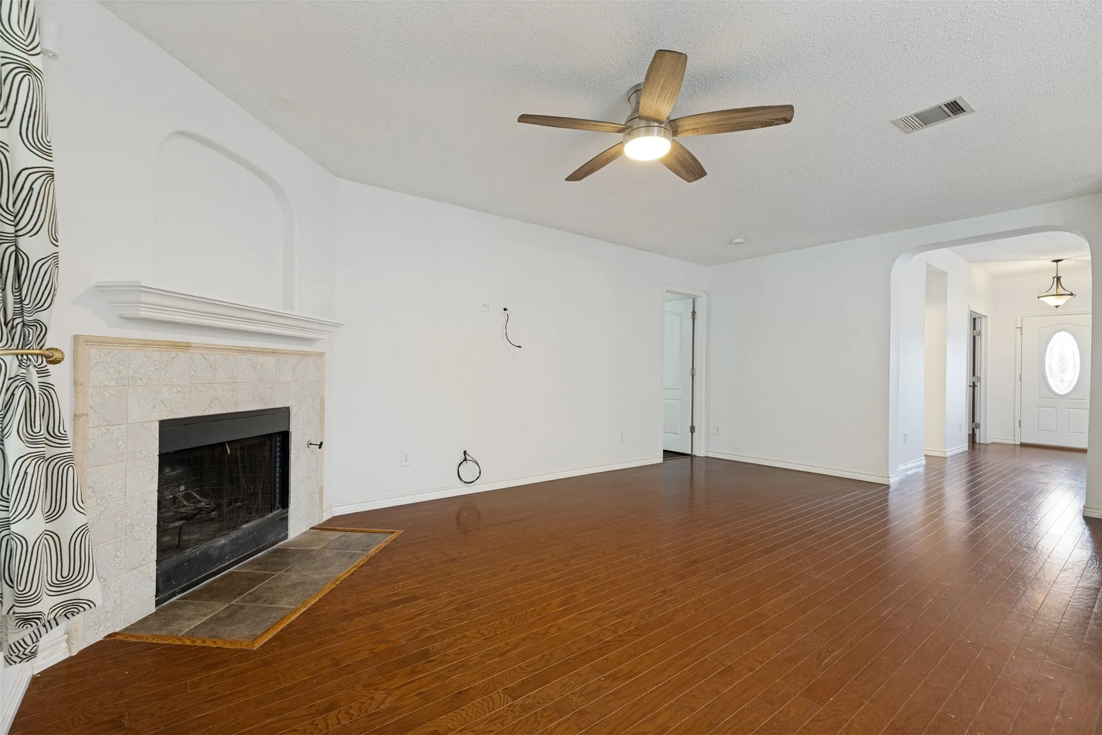 Living area featuring wood-finish flooring, a fireplace with a tiled surround and hearth, a ceiling fan with integrated lighting, and an arched entryway