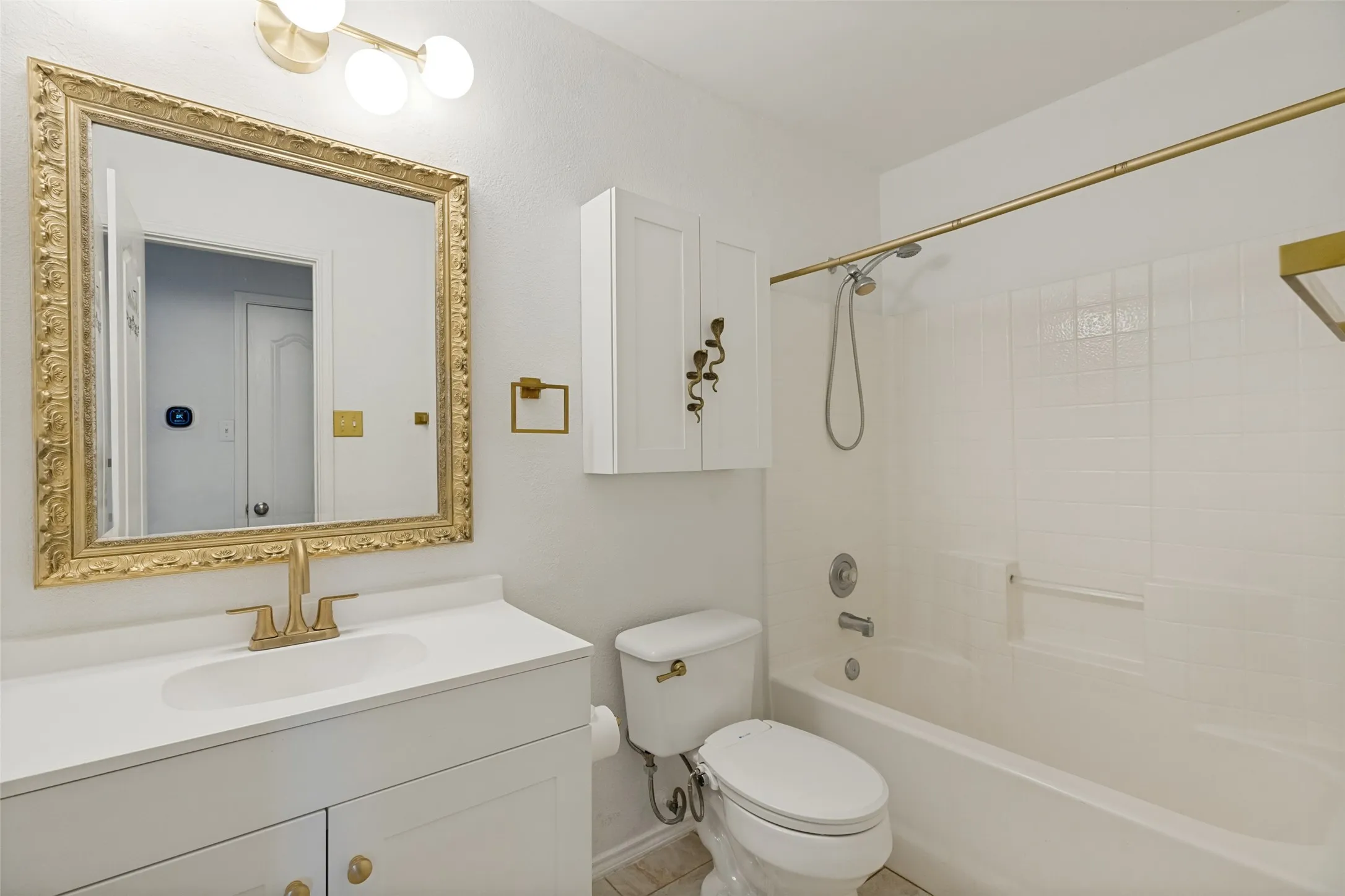 White vanity with integrated sink, ornate gold-framed mirror, three-light vanity fixture, white subway-tiled tub surround, and gold-finish hardware