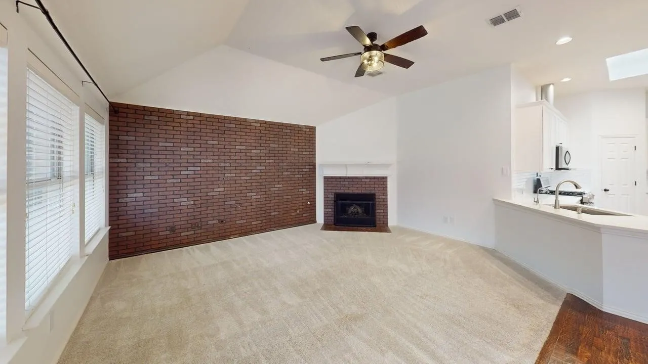 Spacious open living area featuring exposed brick fireplace wall, wood-finish flooring, ceiling fan with integrated lighting, recessed ceiling lights, and direct access to a full bathroom with white cabinetry and sink