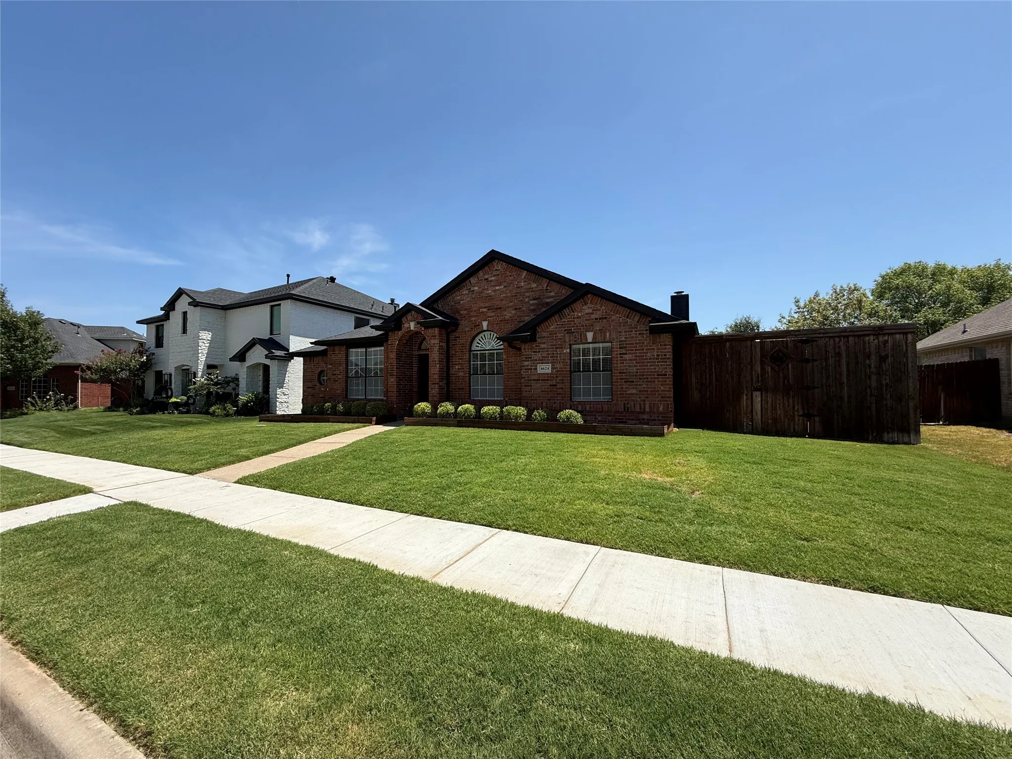 Brick Colonial home showcasing a manicured front lawn with mature landscaping, concrete driveway, and established perimeter fencing