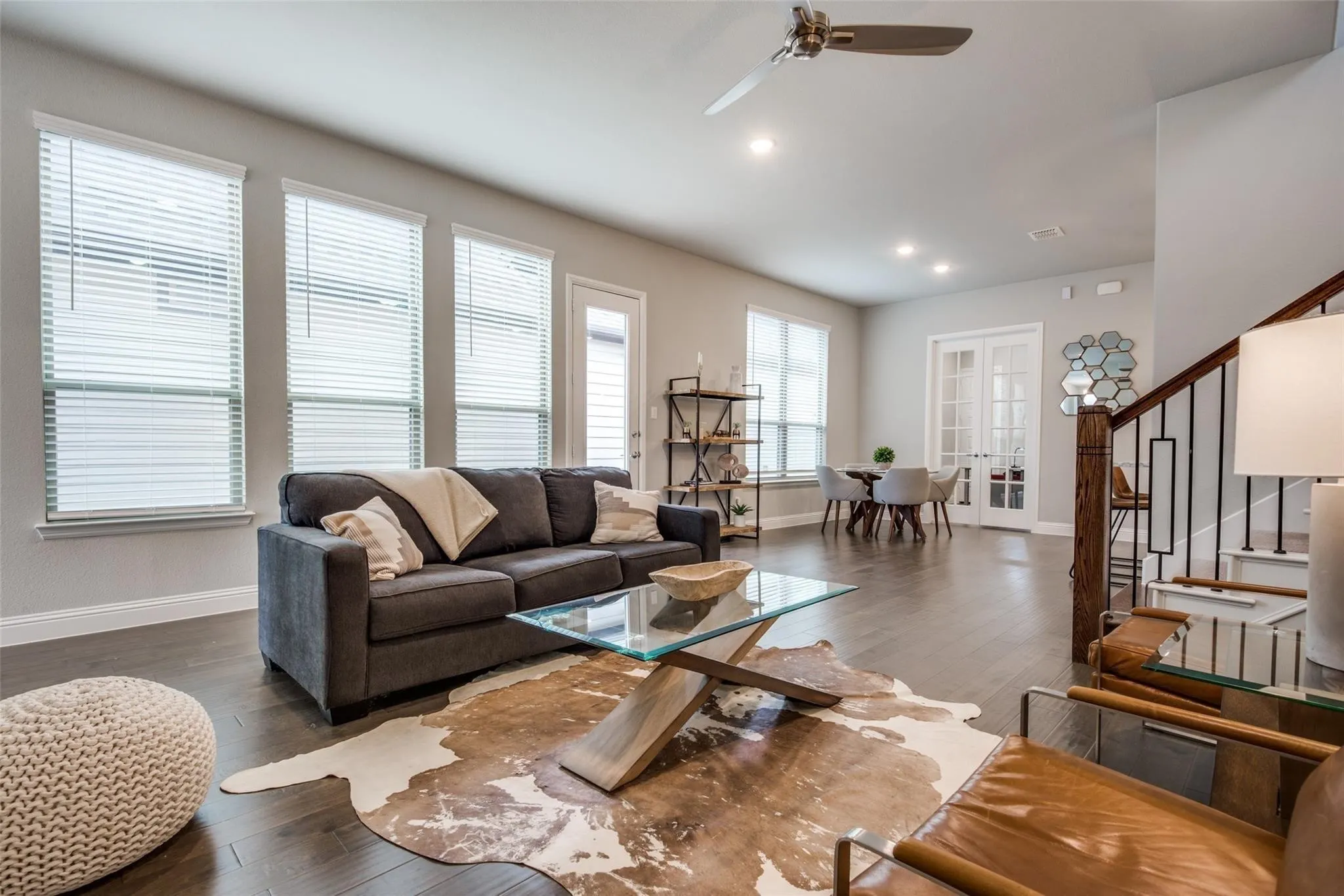 Bright open-concept living space featuring expansive windows with plantation shutters, gleaming wood-finish flooring, and soaring ceilings with ceiling fan