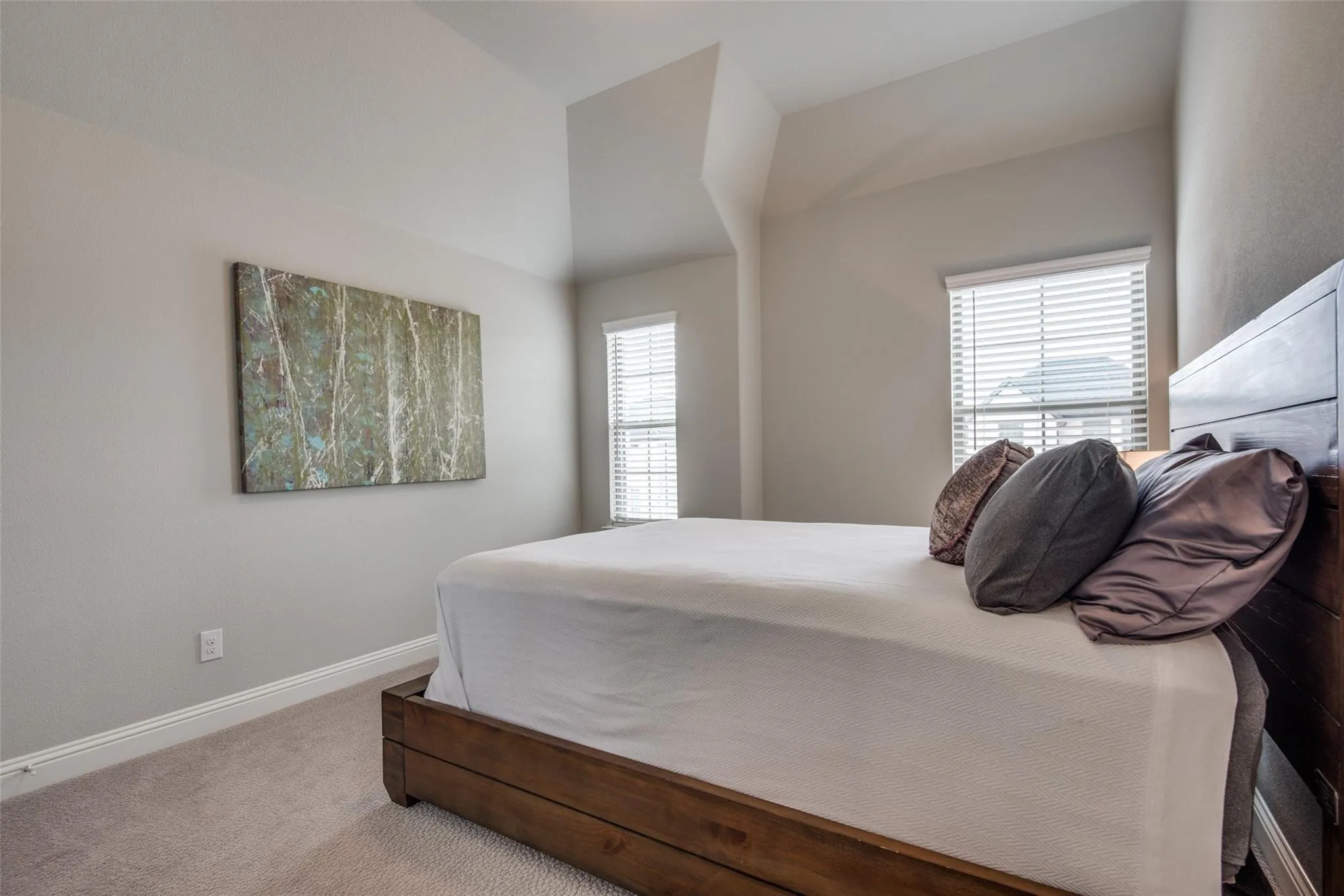 Bright, light-filled bedroom showcasing a wood-finish platform bed frame, neutral carpet flooring, and expansive windows with plantation shutters providing natural light and privacy