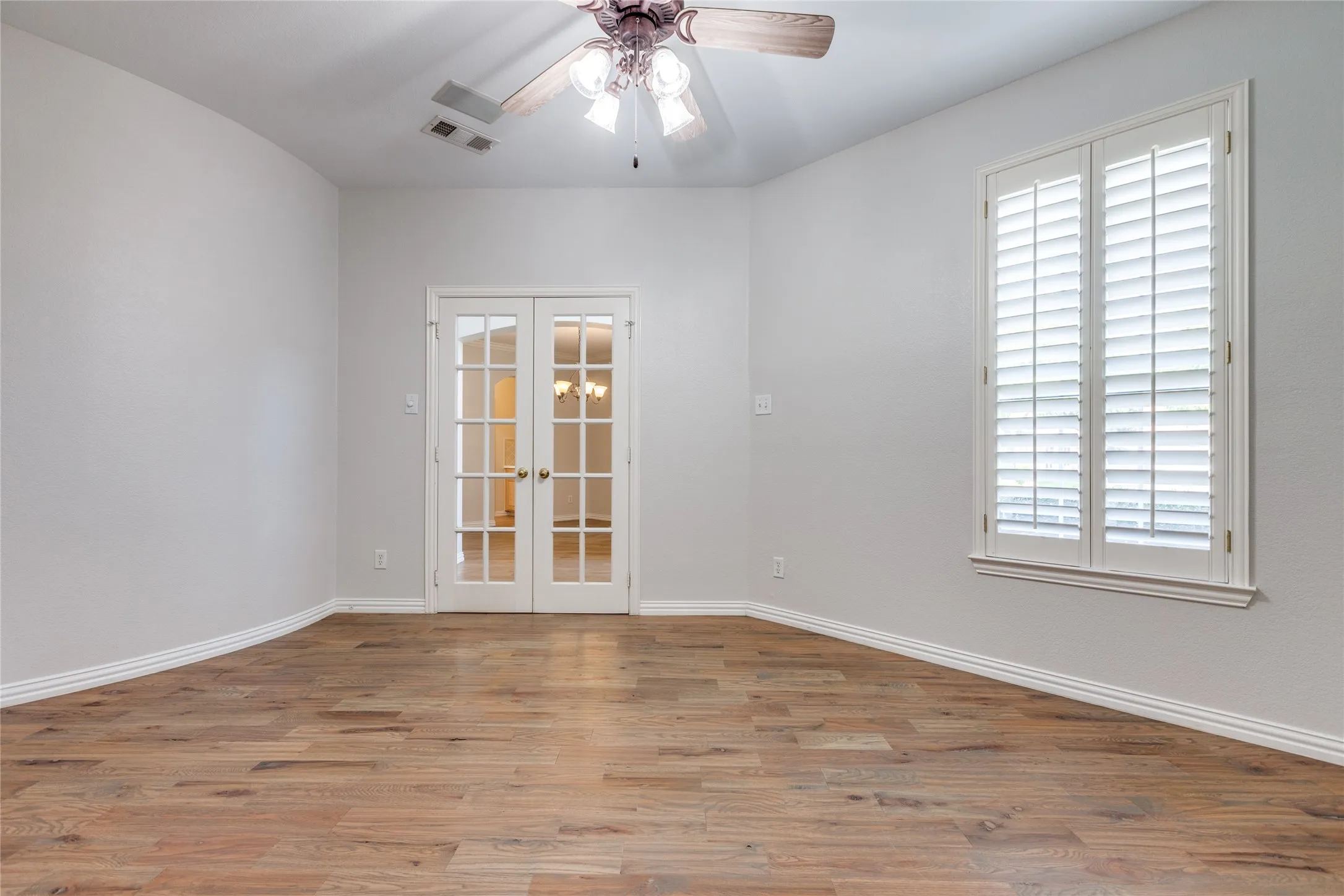 Spacious room showcasing warm wood-finish flooring, white painted walls, and abundant natural light from dual shuttered windows