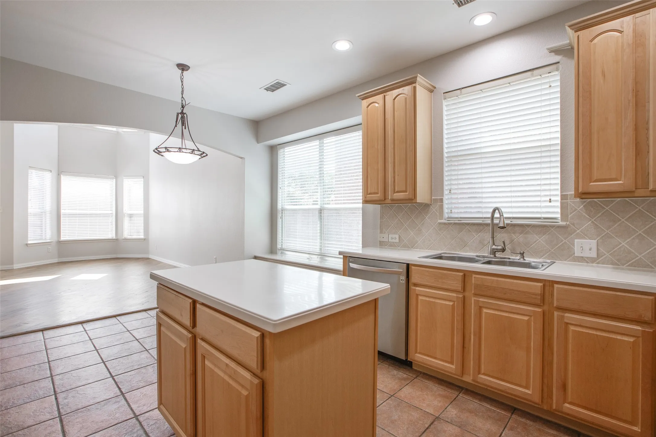 Spacious kitchen featuring warm wood cabinetry with raised-panel doors, a large center island with white quartz countertop, stainless steel sink with faucet, tile flooring, and abundant natural light from multiple windows with blinds