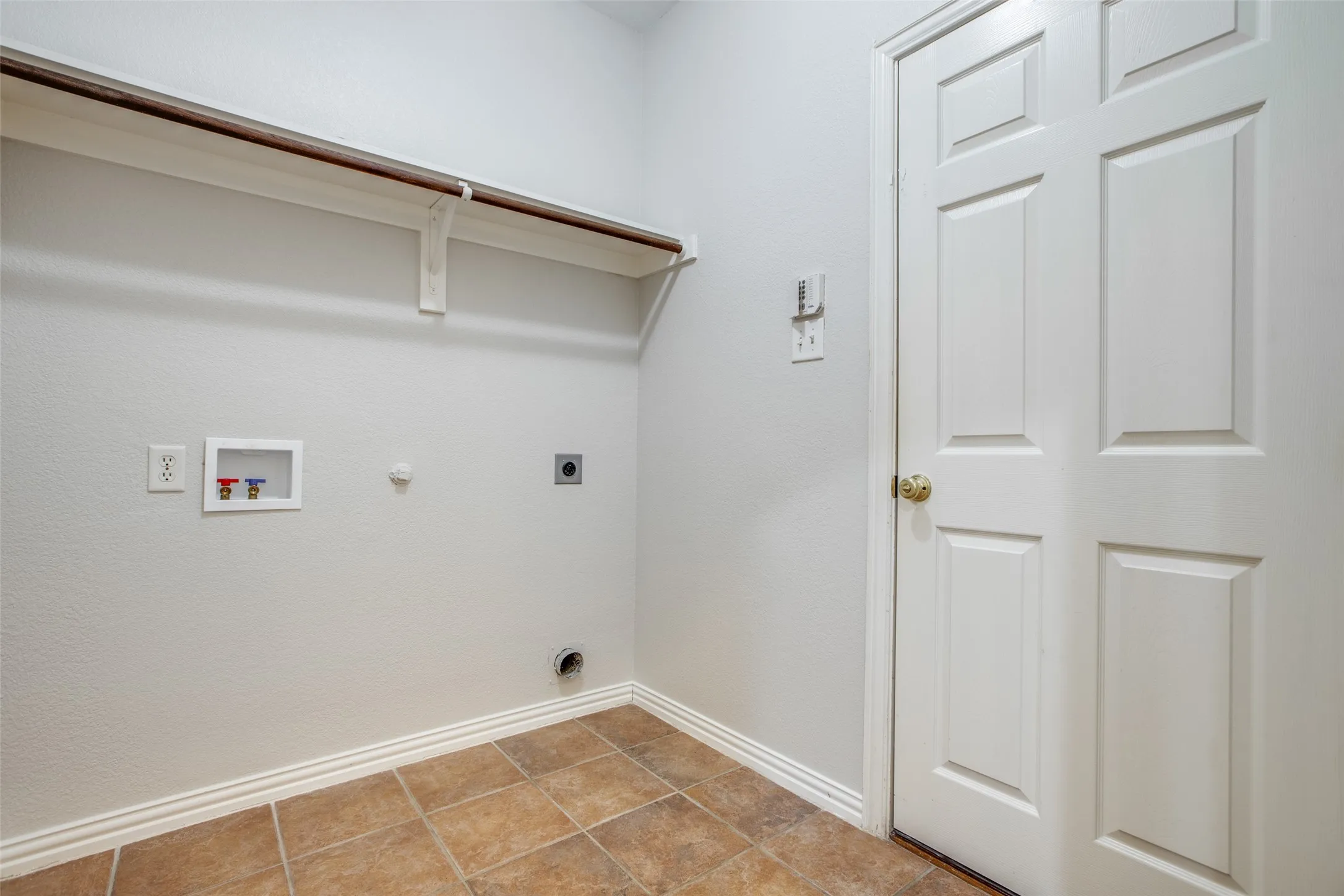 Utility room featuring ceramic tile flooring, wall-mounted shelving with bracket support, electrical outlets, and a six-panel white entry door with brass hardware