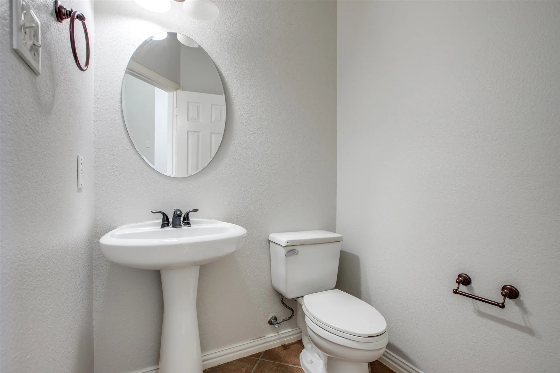 Powder room featuring a pedestal sink with oil-rubbed bronze faucet and hardware, a round mirror with metal frame, a toilet with chrome tank lever, and wood-finish flooring