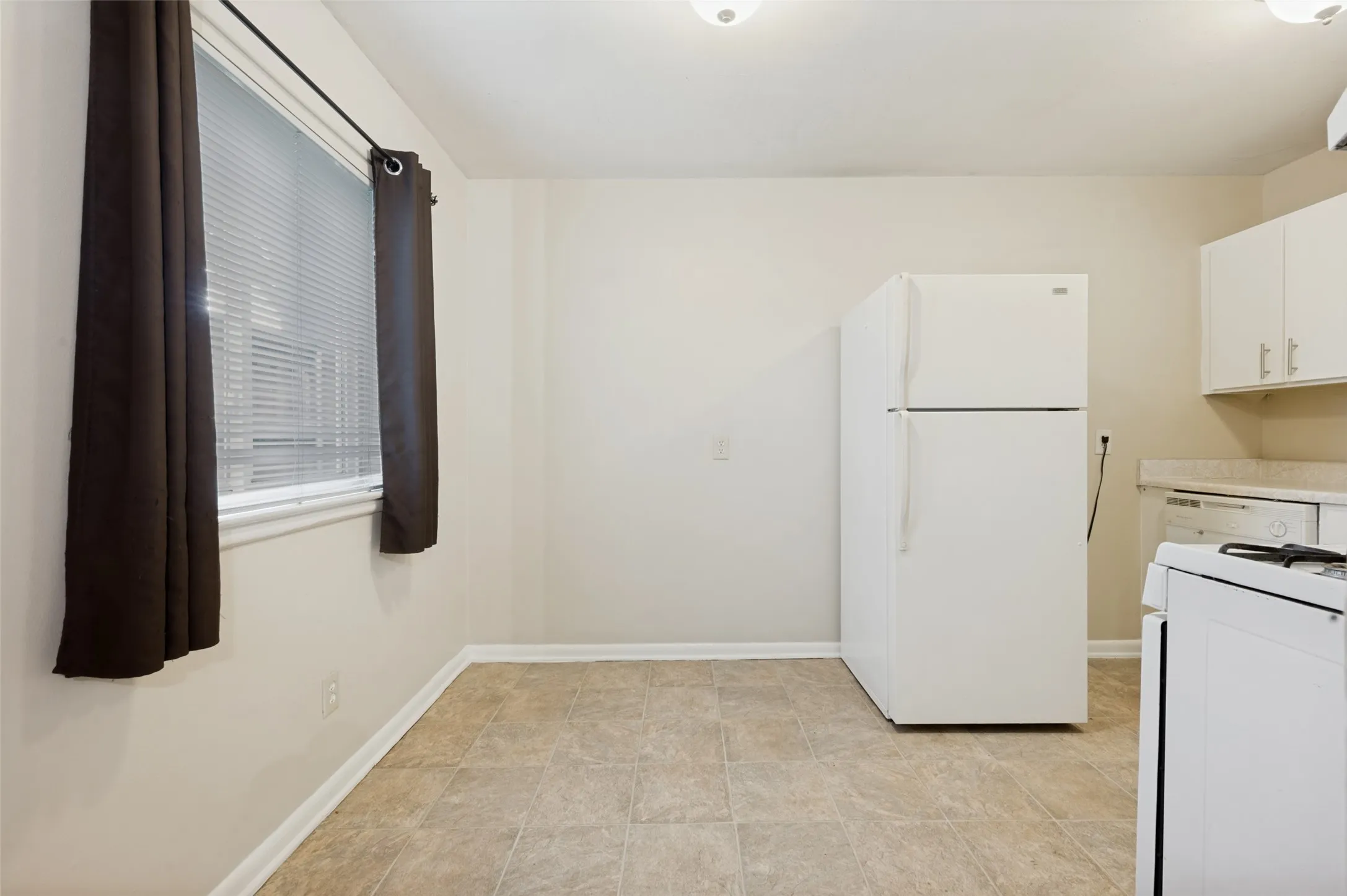 Bright, functional kitchen featuring white appliances including a refrigerator-freezer and electric range, neutral tile flooring, ample cabinet storage, and a window with natural light