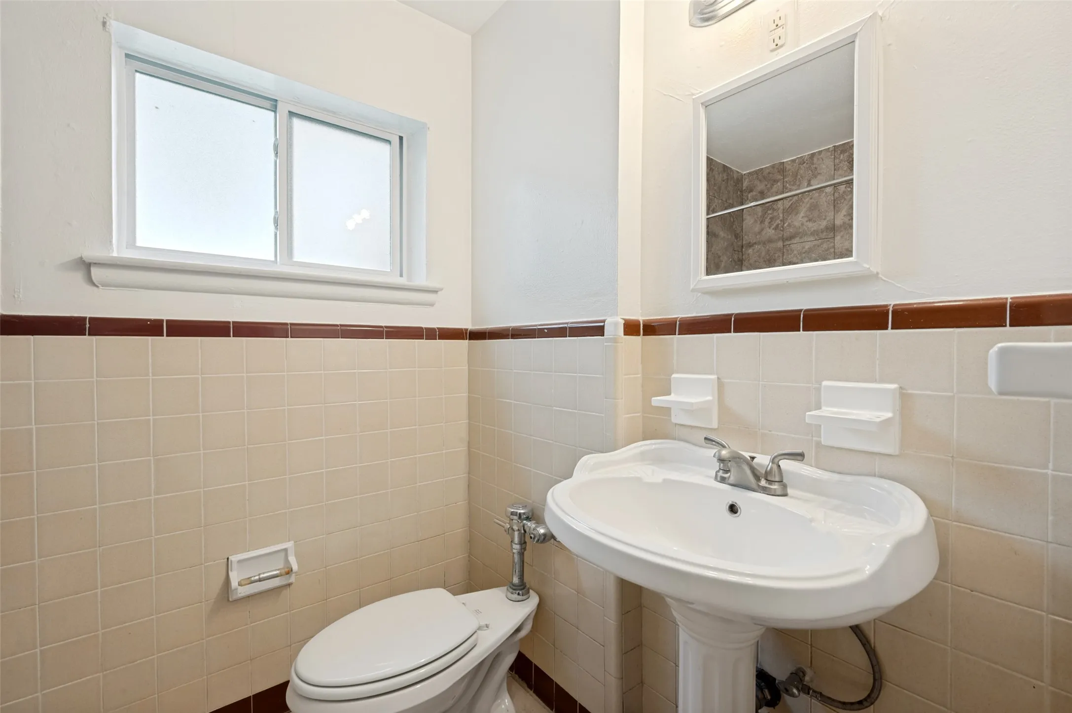 Bright, functional bathroom featuring a pedestal sink with chrome faucet, ceramic tile walls with decorative tile trim, toilet with white seat, and dual windows providing natural light