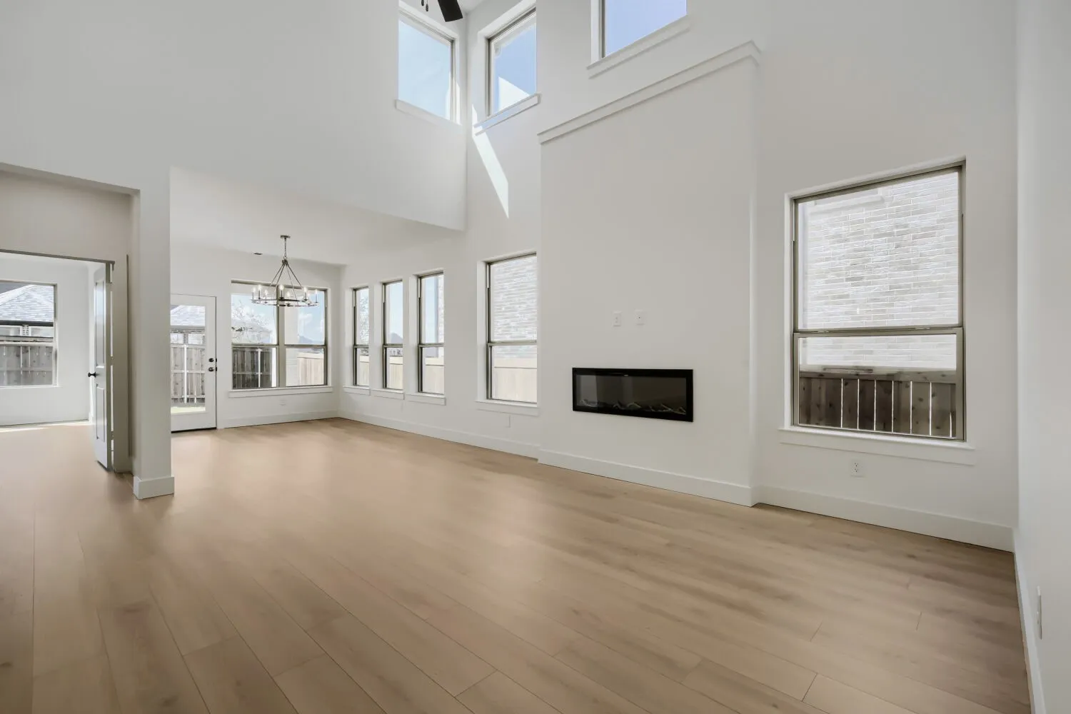 Two-story great room with light wood-finish flooring, multiple windows, a linear electric fireplace, and a contemporary chandelier