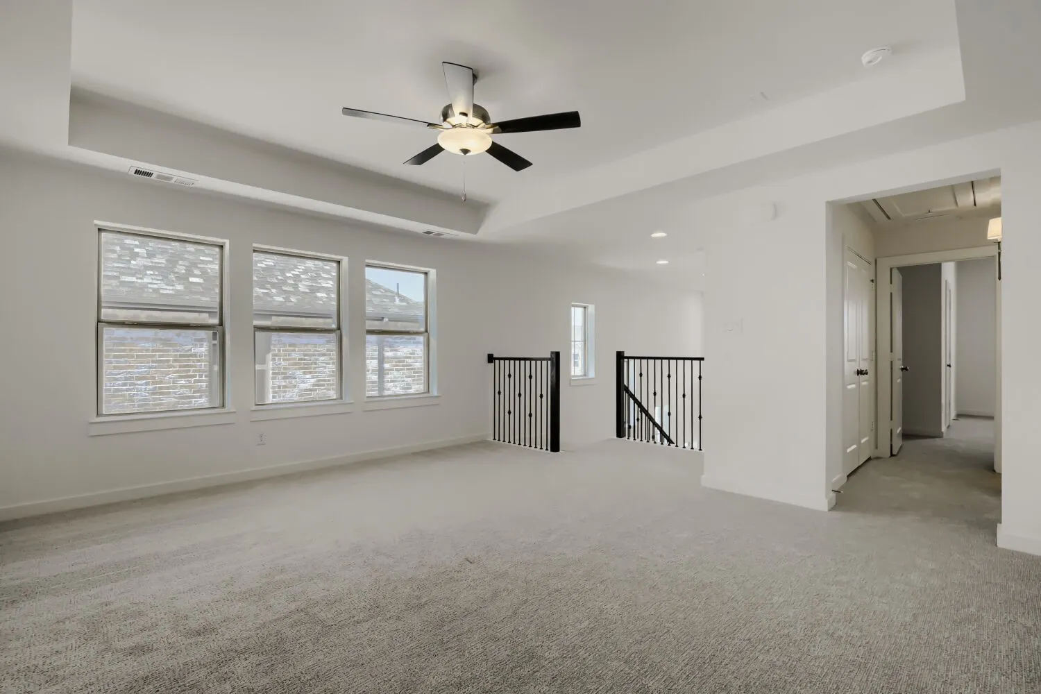 Carpeted loft featuring a tray ceiling, ceiling fan, three windows, and wrought iron balusters
