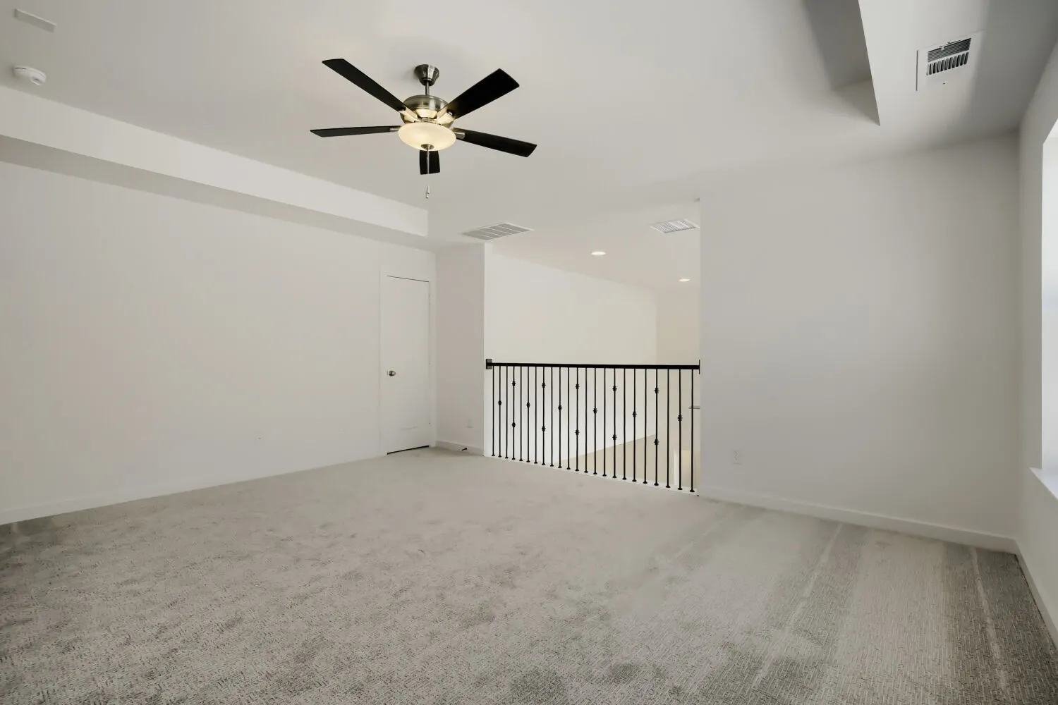 Spacious room with light gray carpet, a ceiling fan with light fixture, and a black wrought iron railing
