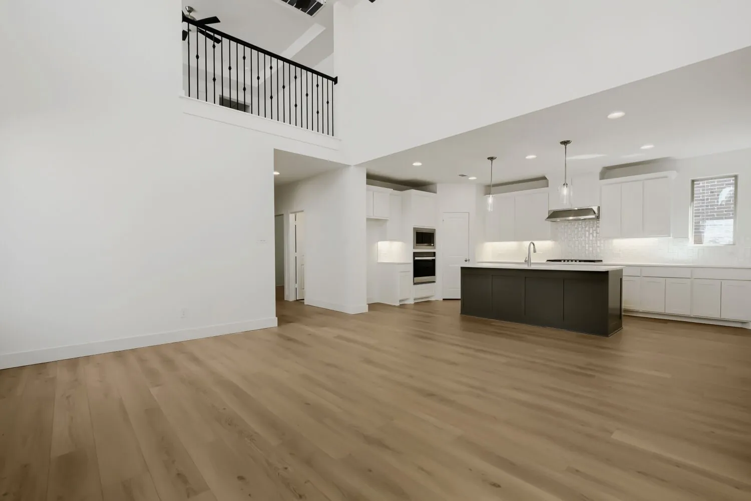 Spacious open-concept living area with wood-finish flooring, a two-story ceiling, and an upper-level balcony with a black iron railing