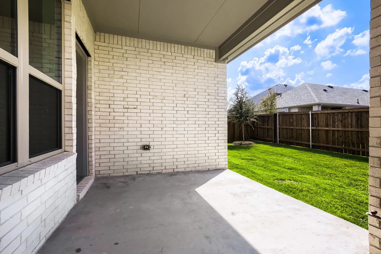 Covered concrete patio with white brick exterior, green lawn, and wood privacy fencing