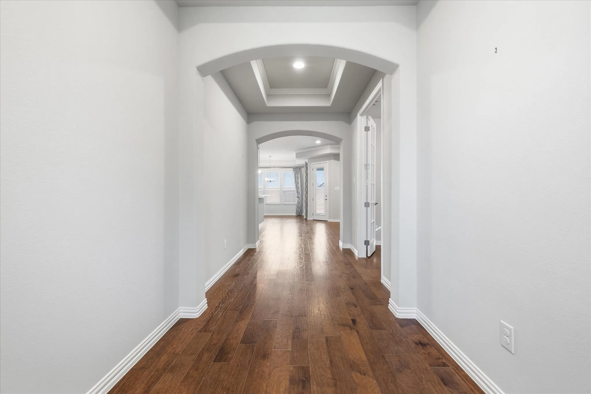 Hallway featuring wood-finish flooring, white baseboards, light grey wall paint, and an arched entry with a coffered ceiling