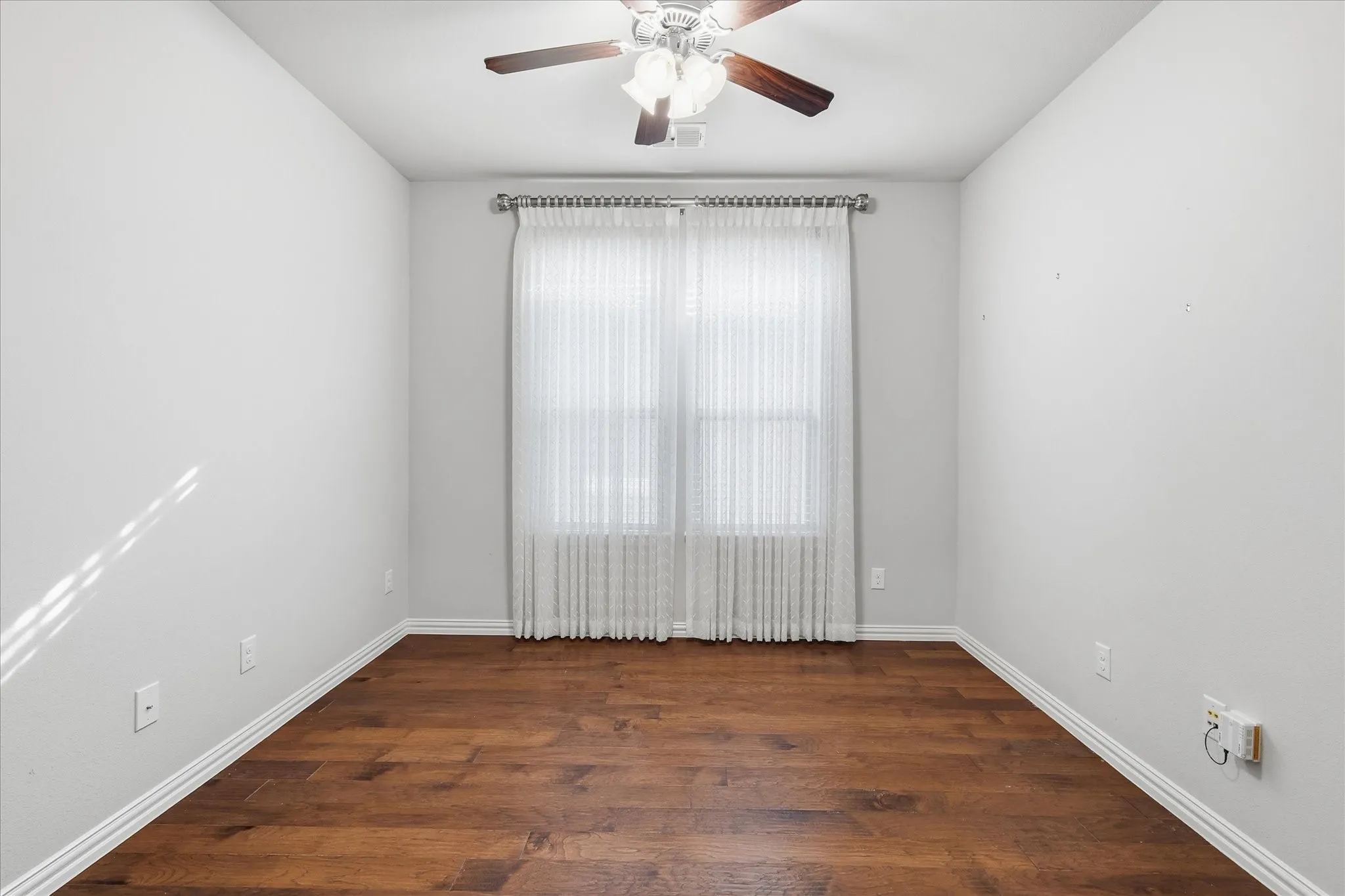 Room featuring wood-finish flooring, a ceiling fan with integrated lighting, and a window with sheer white curtains