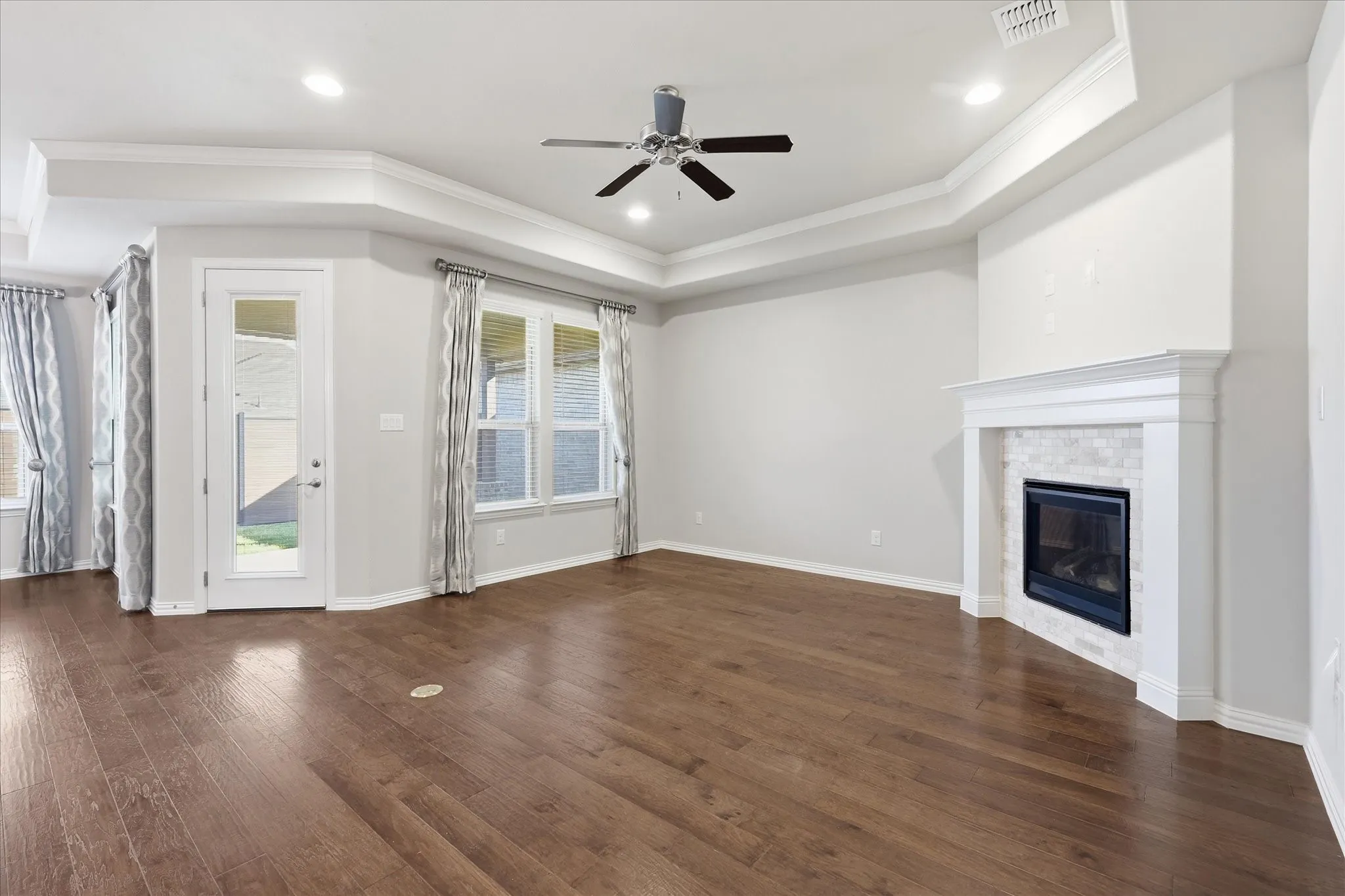 Spacious living area featuring warm wood-finish flooring, coffered ceiling with recessed lighting and ceiling fan, gas fireplace with white surround and mantel, French doors with transom windows and thermal-lined curtains, and direct access to exterior spaces