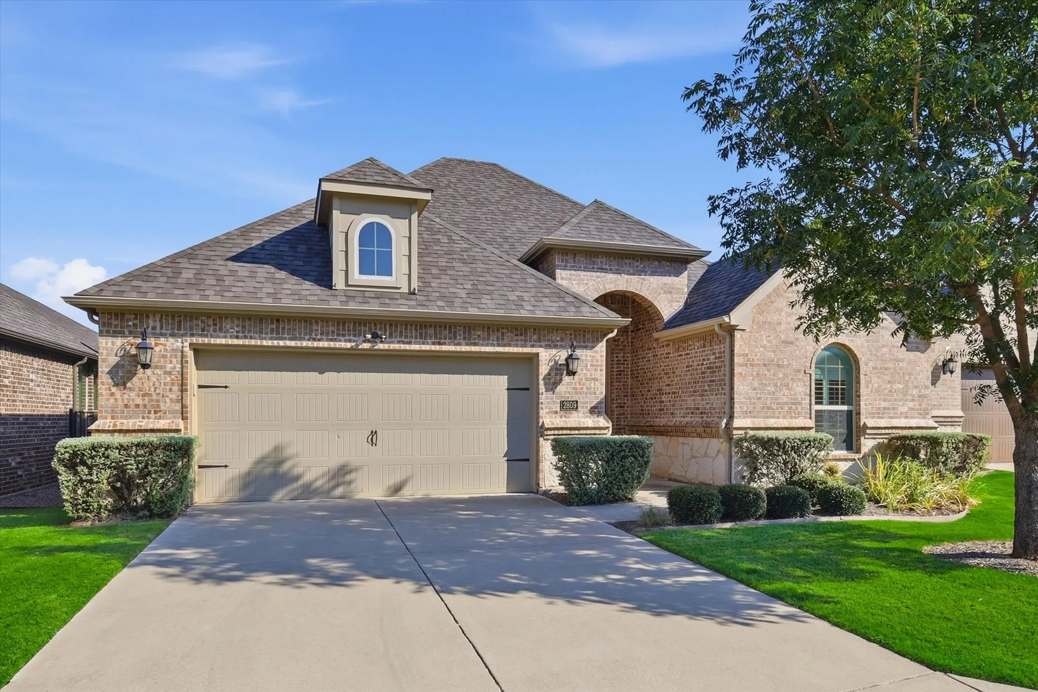 Brick exterior featuring a two-car garage, dormer window, arched entry, and established landscaping