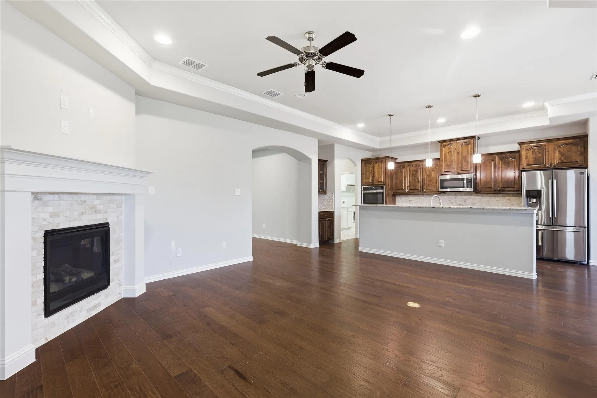 Spacious open-concept living area featuring wood-finish flooring, a corner fireplace with a tiled surround, and tray ceilings with recessed lighting