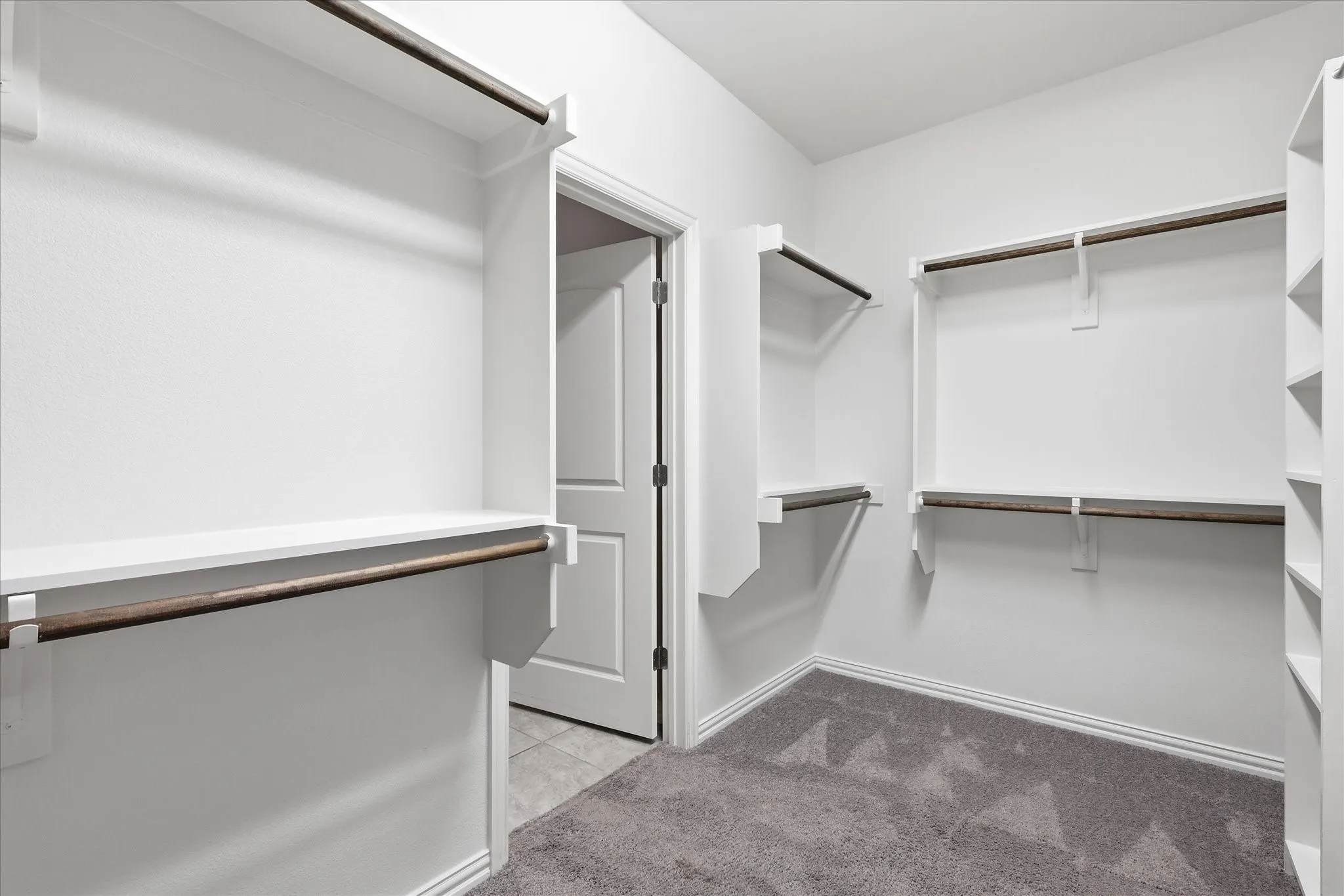 Spacious walk-in closet featuring white built-in shelving, dark wood-finish hanging rods, and light gray patterned carpeting