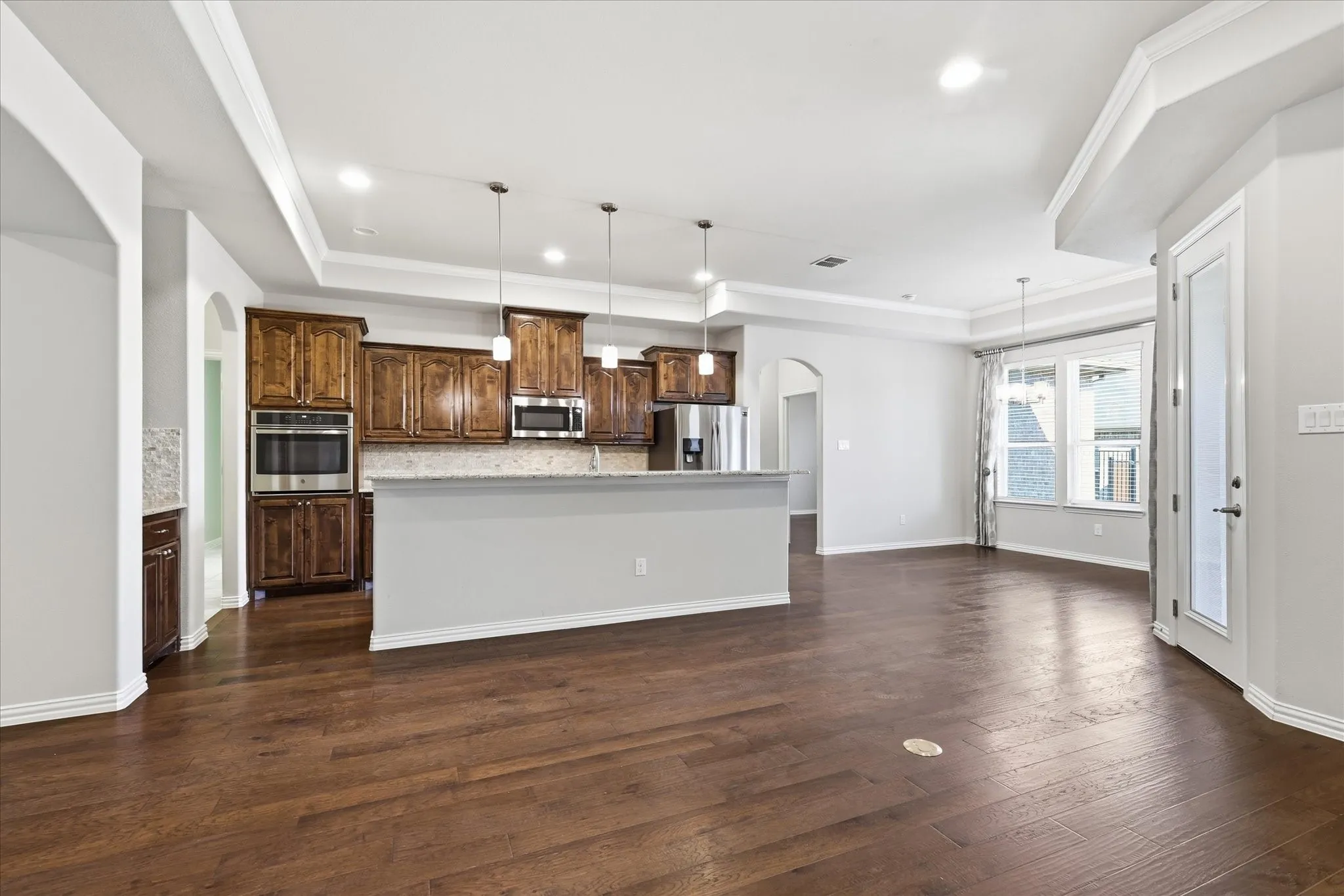 Open-concept living area featuring rich wood-finish flooring, a tray ceiling with recessed lighting, and a kitchen island with pendant lighting