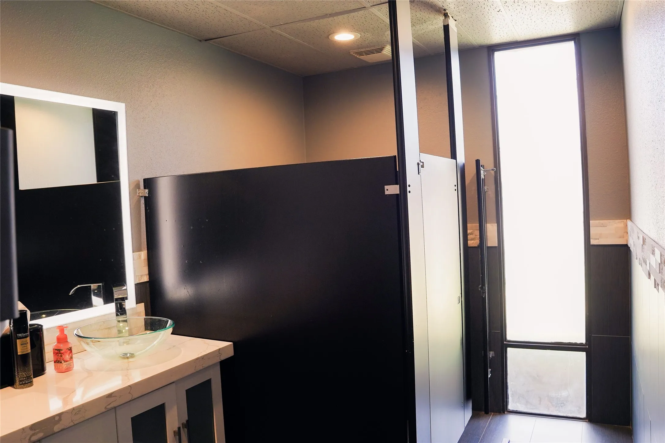 Restroom with privacy partitions, a vanity featuring a vessel sink, and an exterior window